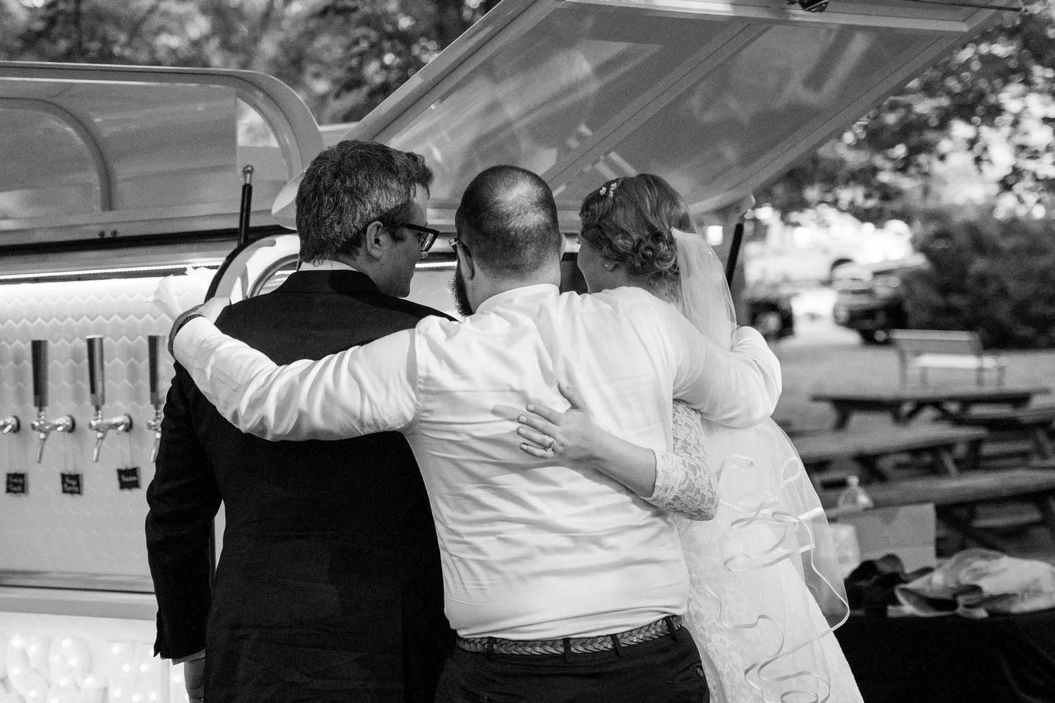 Black and white photo of three people in a warm embrace beside an open vehicle trunk at an outdoor wedding celebration.
