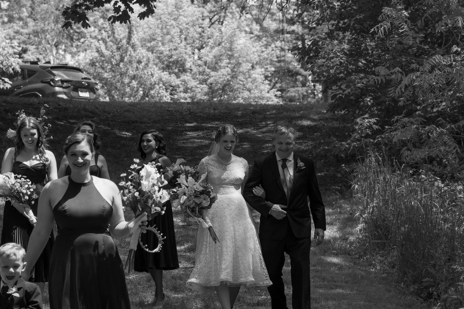 A black and white photo of a bride in a short dress walking with an older man, followed by bridesmaids carrying bouquets.