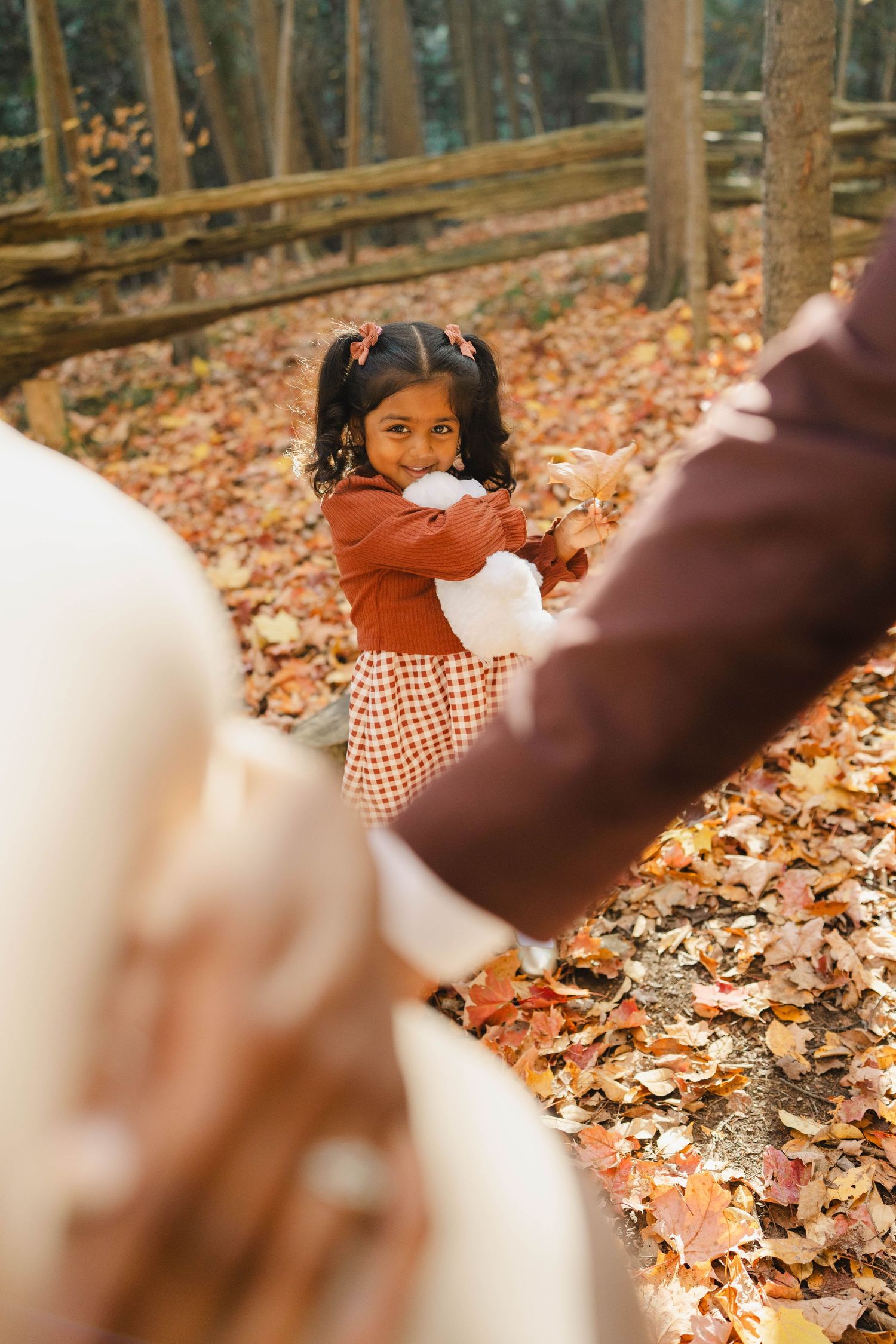 Happy toddler girl holding a white teddy bear stands among colorful fall leaves while her parents hold hands in the foreground.