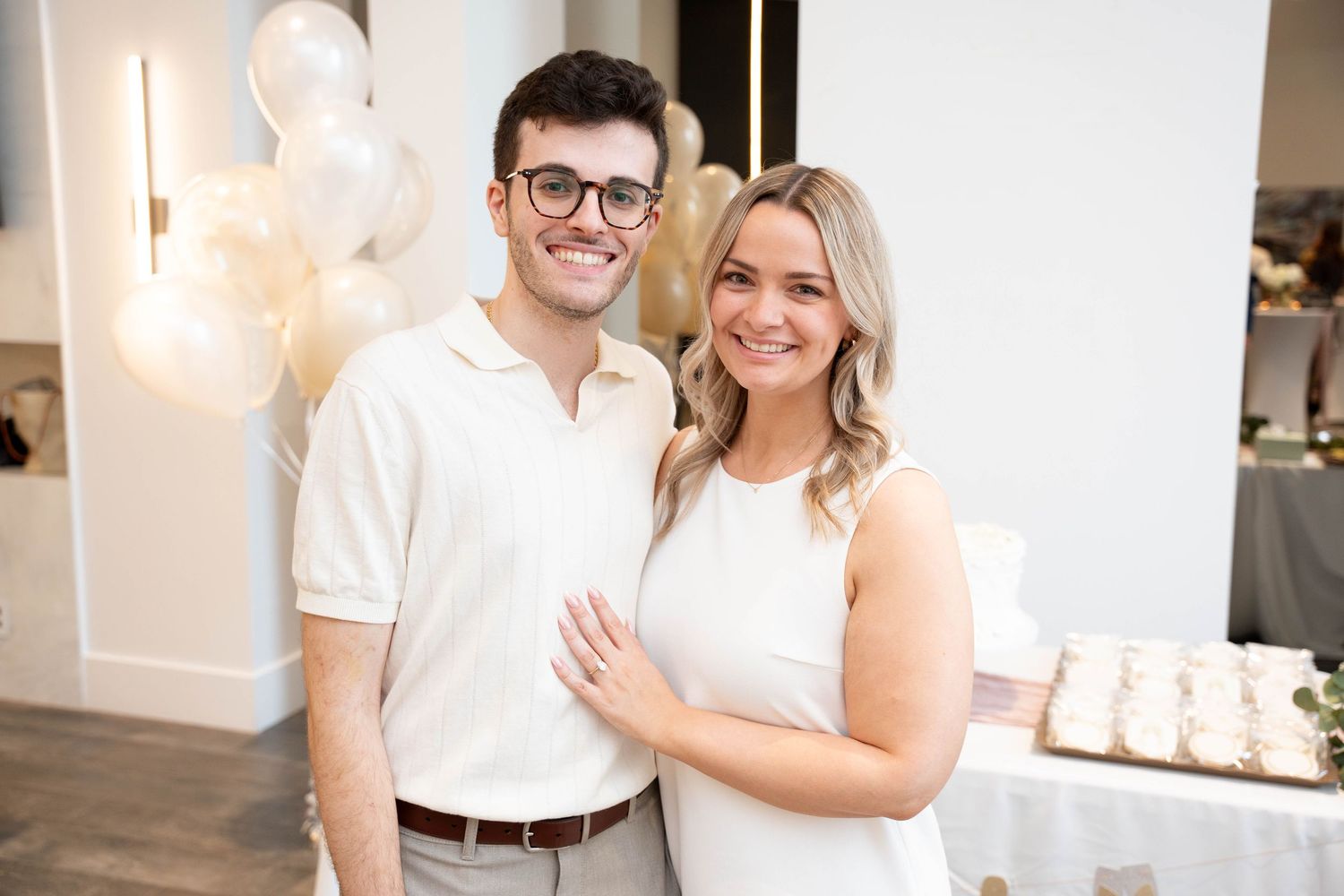Smiling couple in white outfits pose together at an engagement party with balloons and a dessert table behind them.