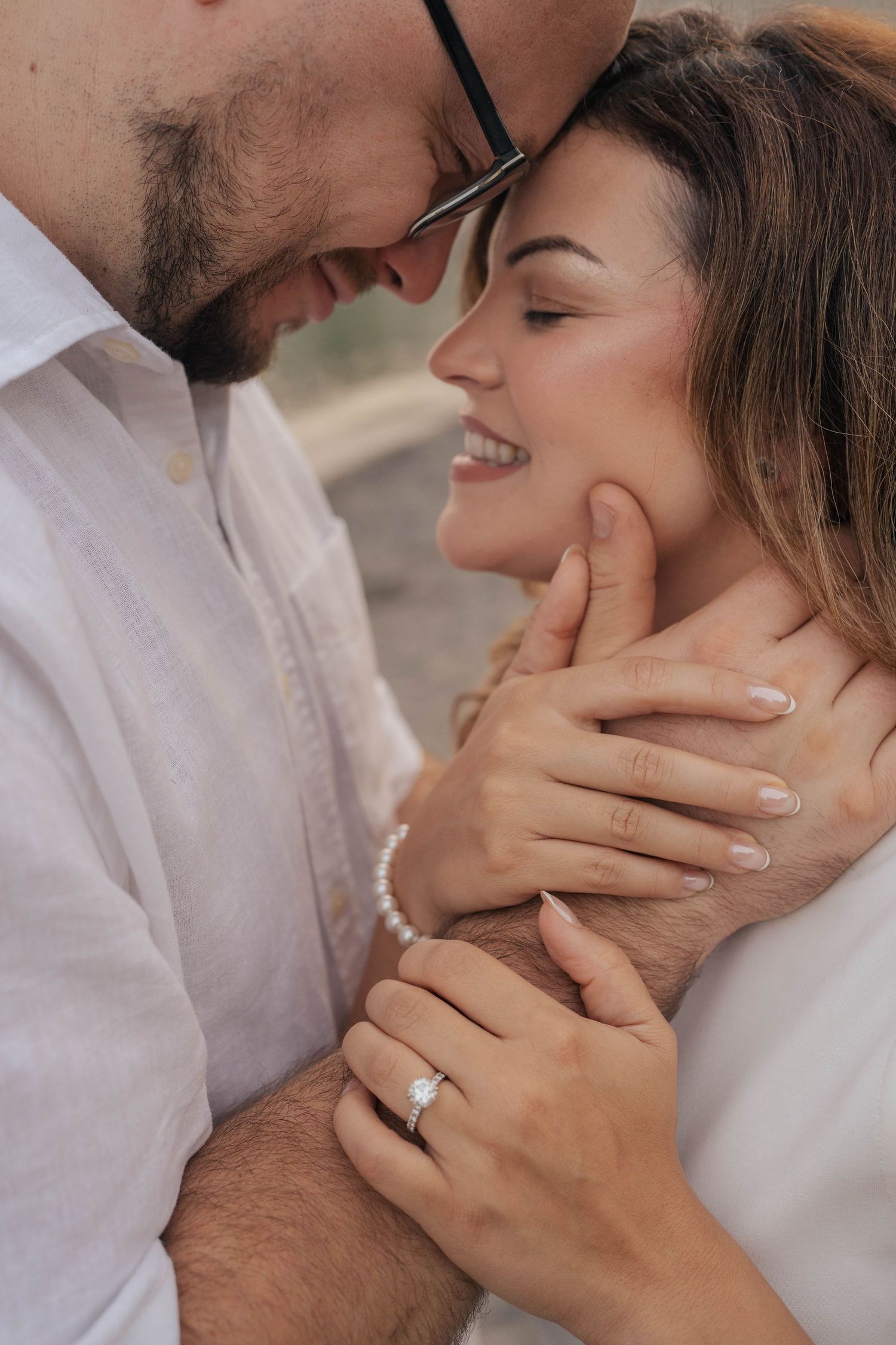Engaged couple touching foreheads and smiling in a tender embrace, her diamond ring and pearl bracelet visible.