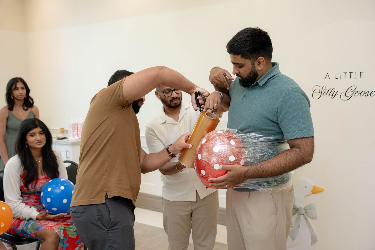 Guests laugh and watch as two men play a baby shower game with a red polka dot balloon beneath a Little Silly Goose sign.