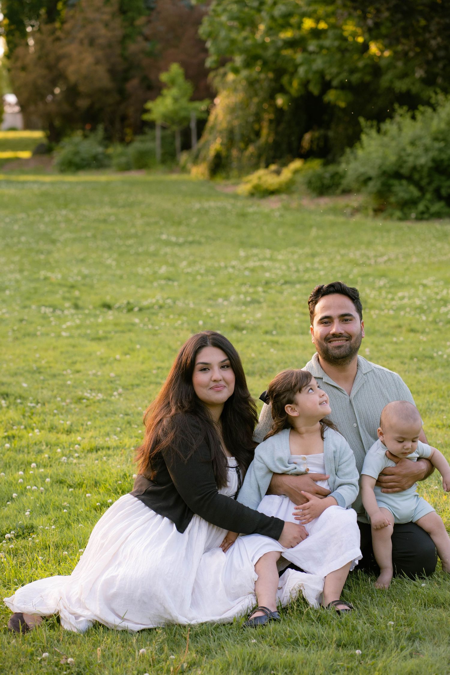 A smiling family of four sits together on green grass in a park, with parents holding their daughter and baby son.