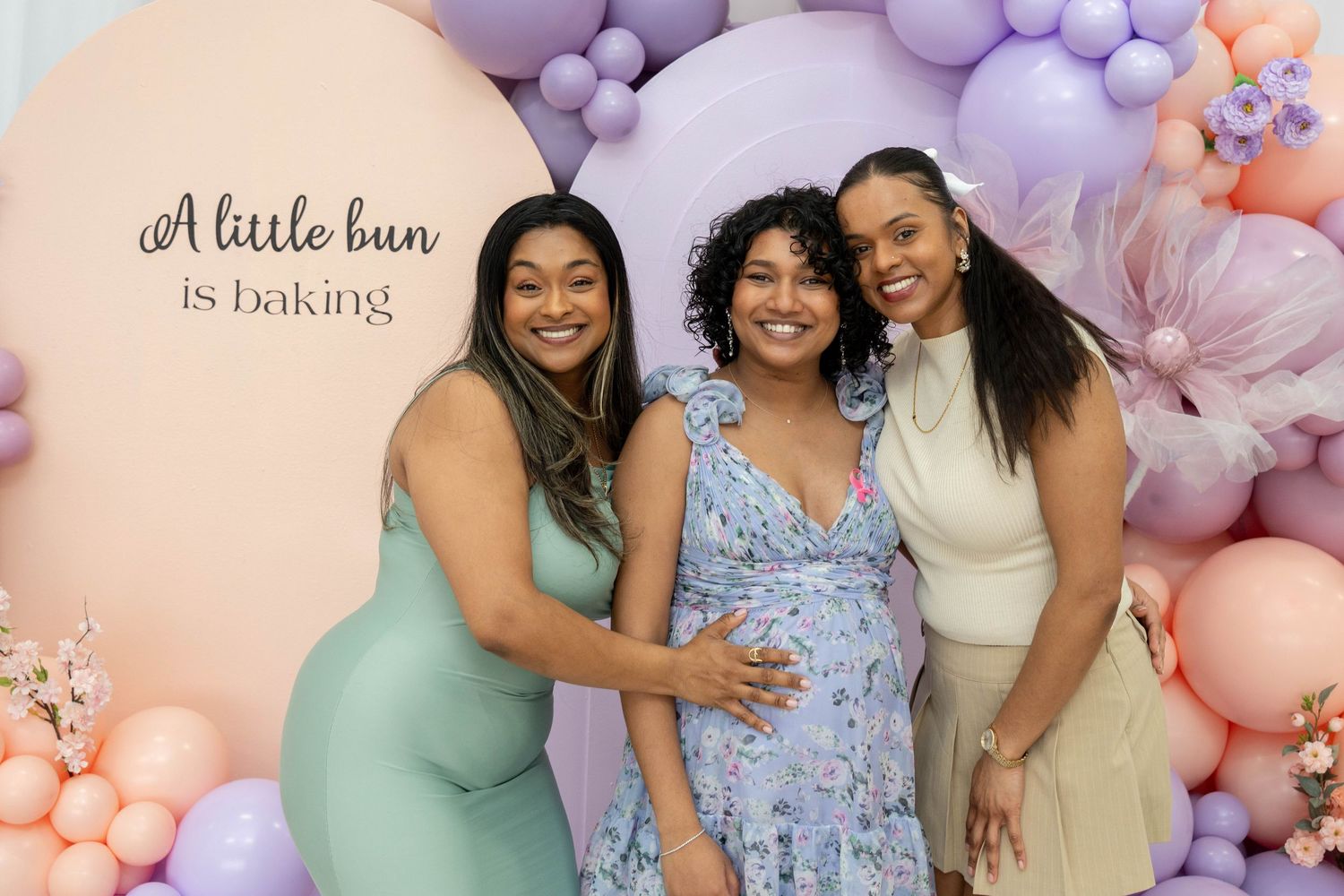 Pregnant woman in a floral dress poses between two friends in front of a 'A little bun is baking' sign and pastel balloon garland.