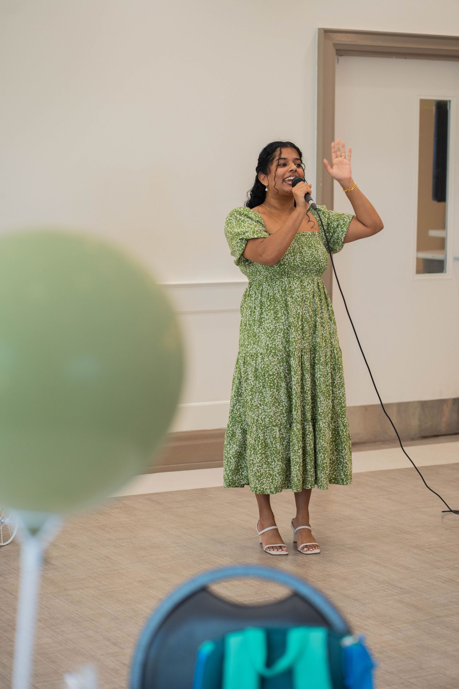 Woman in a green floral dress sings into a microphone and waves at a baby shower, with a green balloon in the foreground.