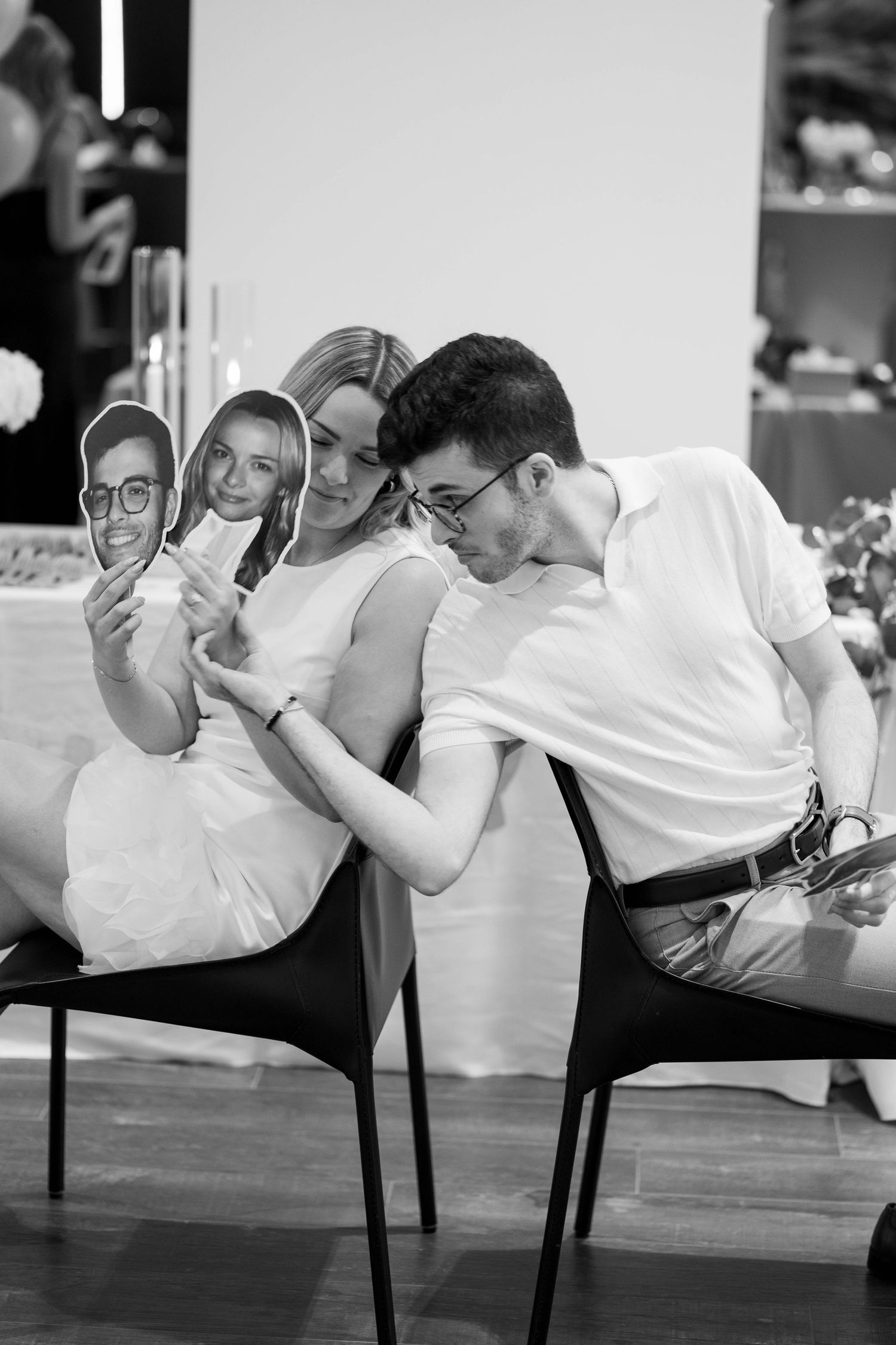In black and white, a smiling couple holds face cutouts on sticks and leans together while seated at an engagement celebration.