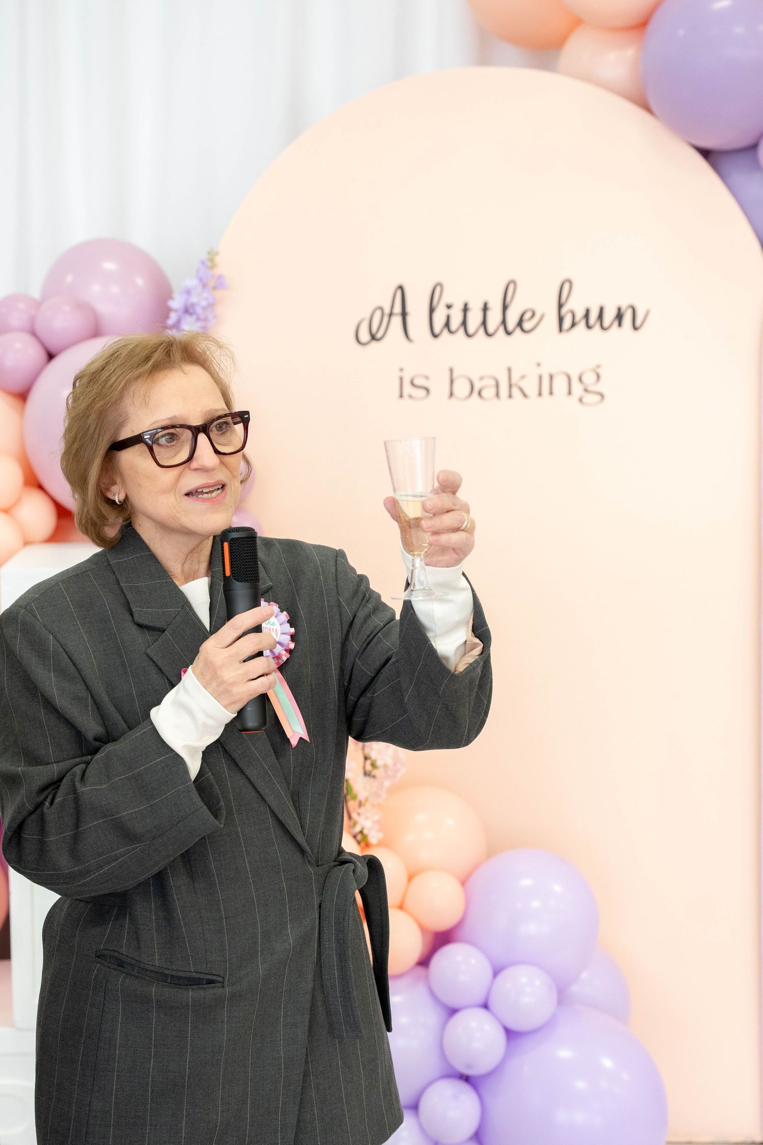 Woman in glasses and a gray blazer raises a champagne glass and speaks into a microphone at a baby shower toast.