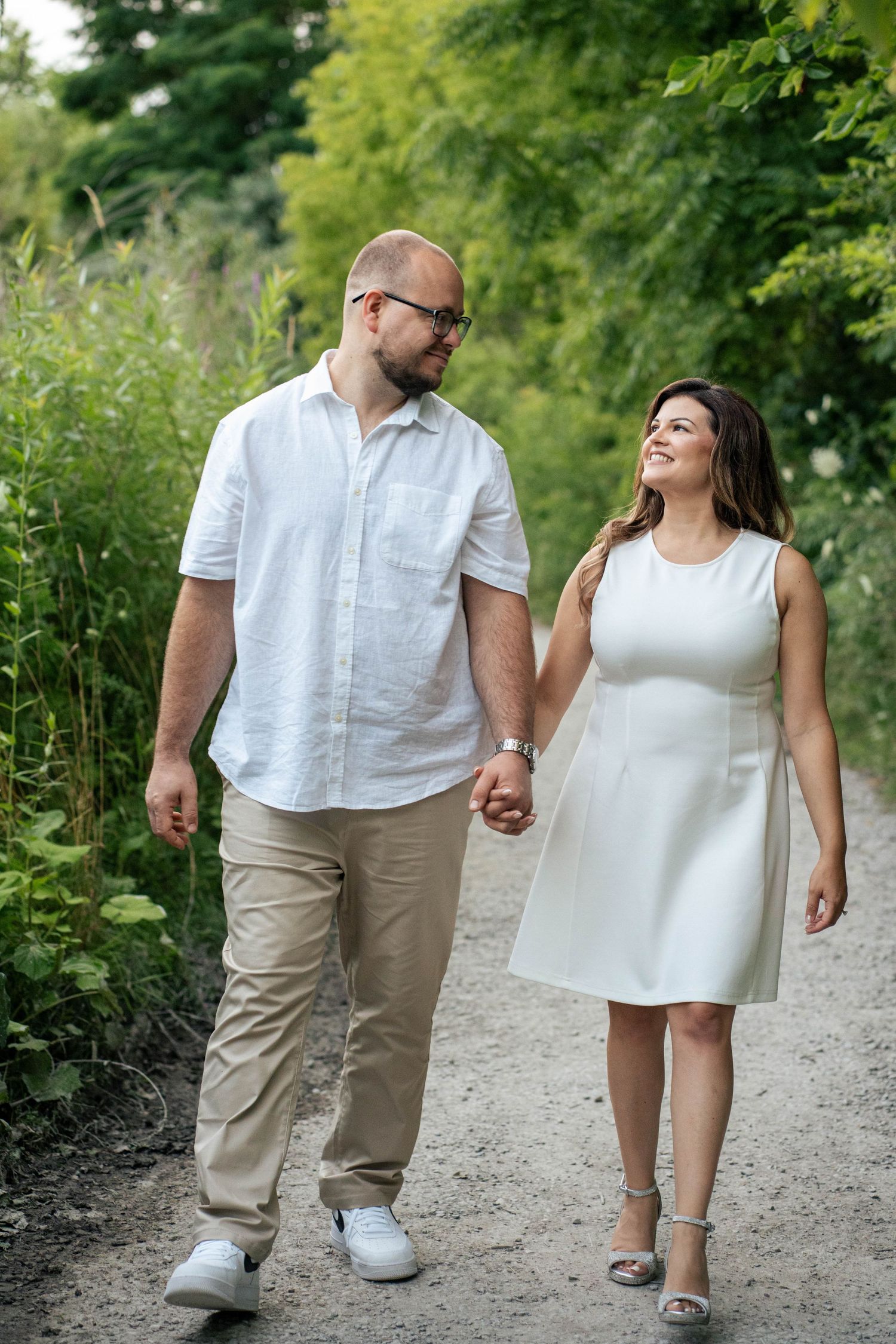 Smiling couple in white and khaki holding hands while walking along a tree-lined gravel path during an engagement photo session.