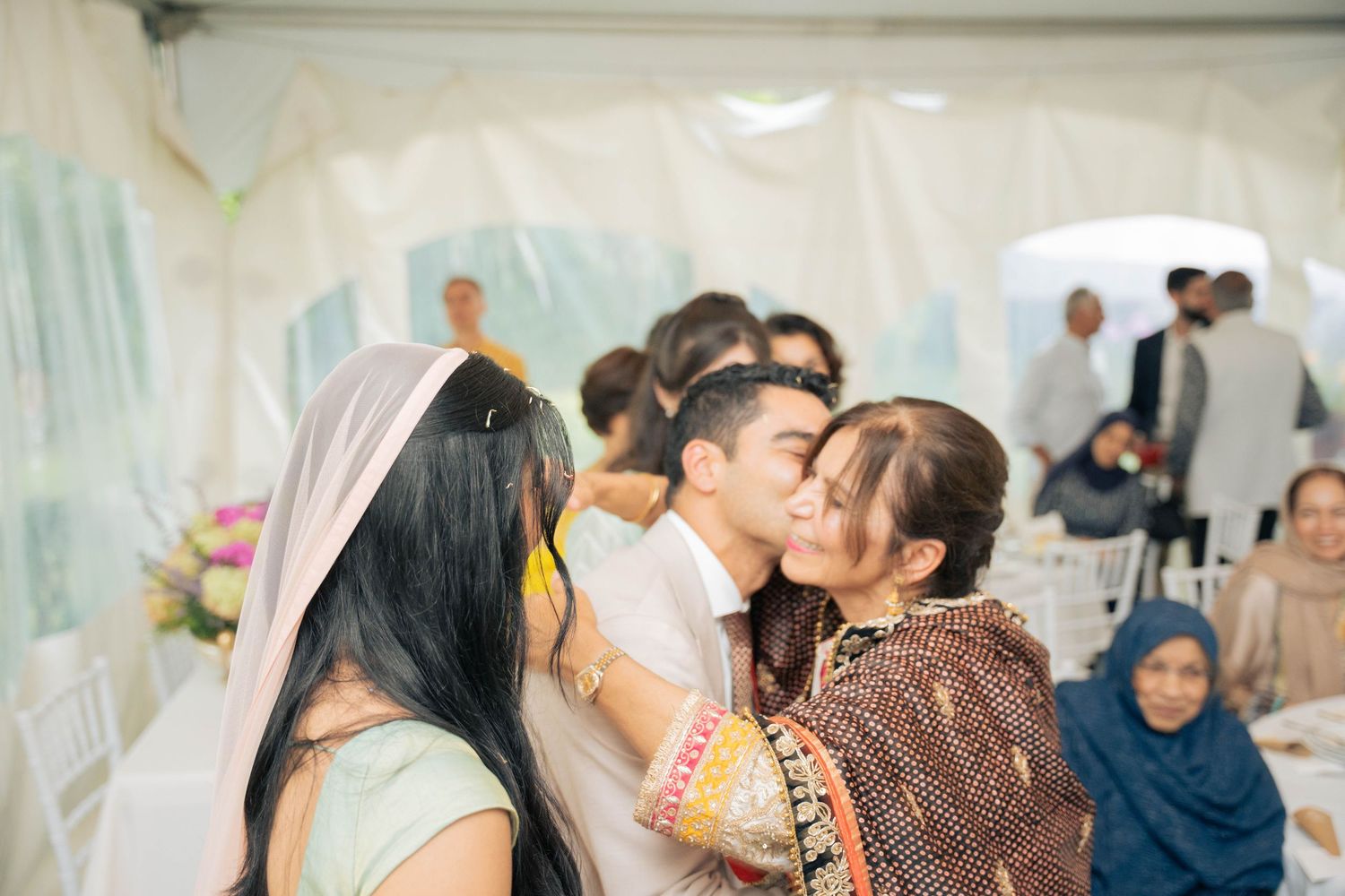 Wedding guests embrace the bride and groom in a joyful, crowded moment under a white tent, with relatives smiling and seated nearby.