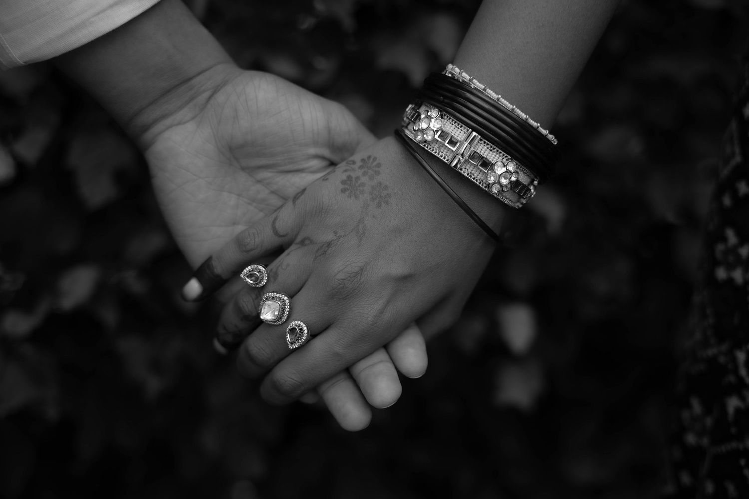 Black and white close-up of two clasped hands, one wearing decorative rings and ornate bangles, against a softly blurred dark background.