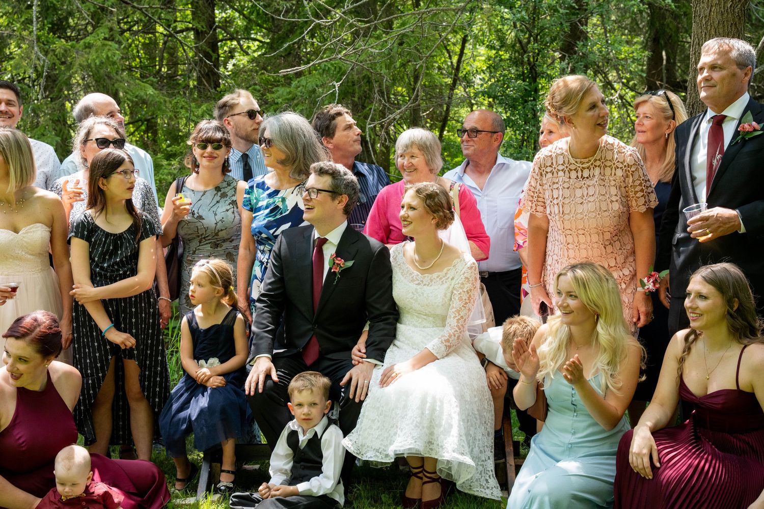 The bride and groom sit on the grass for a group photo surrounded by smiling guests, including children, in a wooded setting.