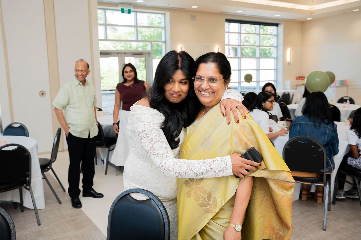 Pregnant woman in white lace hugs an older woman in a yellow sari while guests mingle at a baby shower in a bright hall.