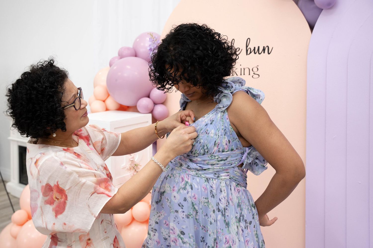 Woman in glasses pins a corsage on an expectant mother in a blue floral dress near pink and purple balloons.