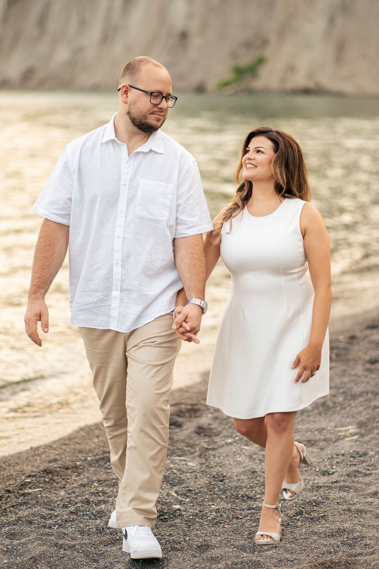 Happy engaged couple walking hand in hand along a pebbly beach with sparkling water and cliffs in the background.