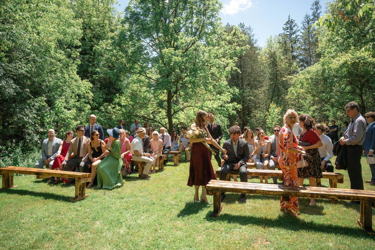 Wedding guests sit on wooden benches on a sunny lawn surrounded by trees as two people walk in front of the ceremony.