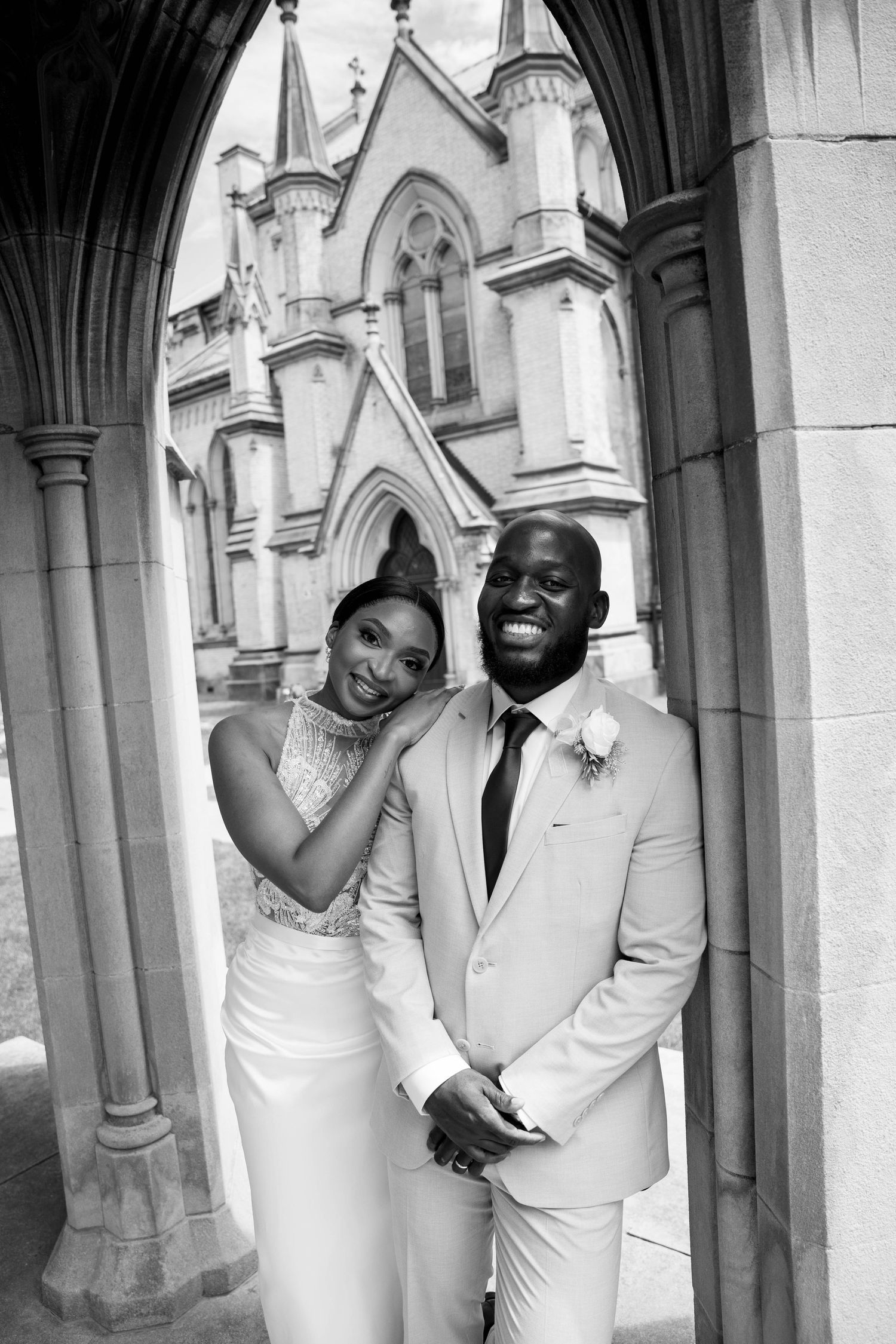 Black and white portrait of a bride leaning affectionately on her smiling groom beneath a stone archway with a Gothic church behind them.