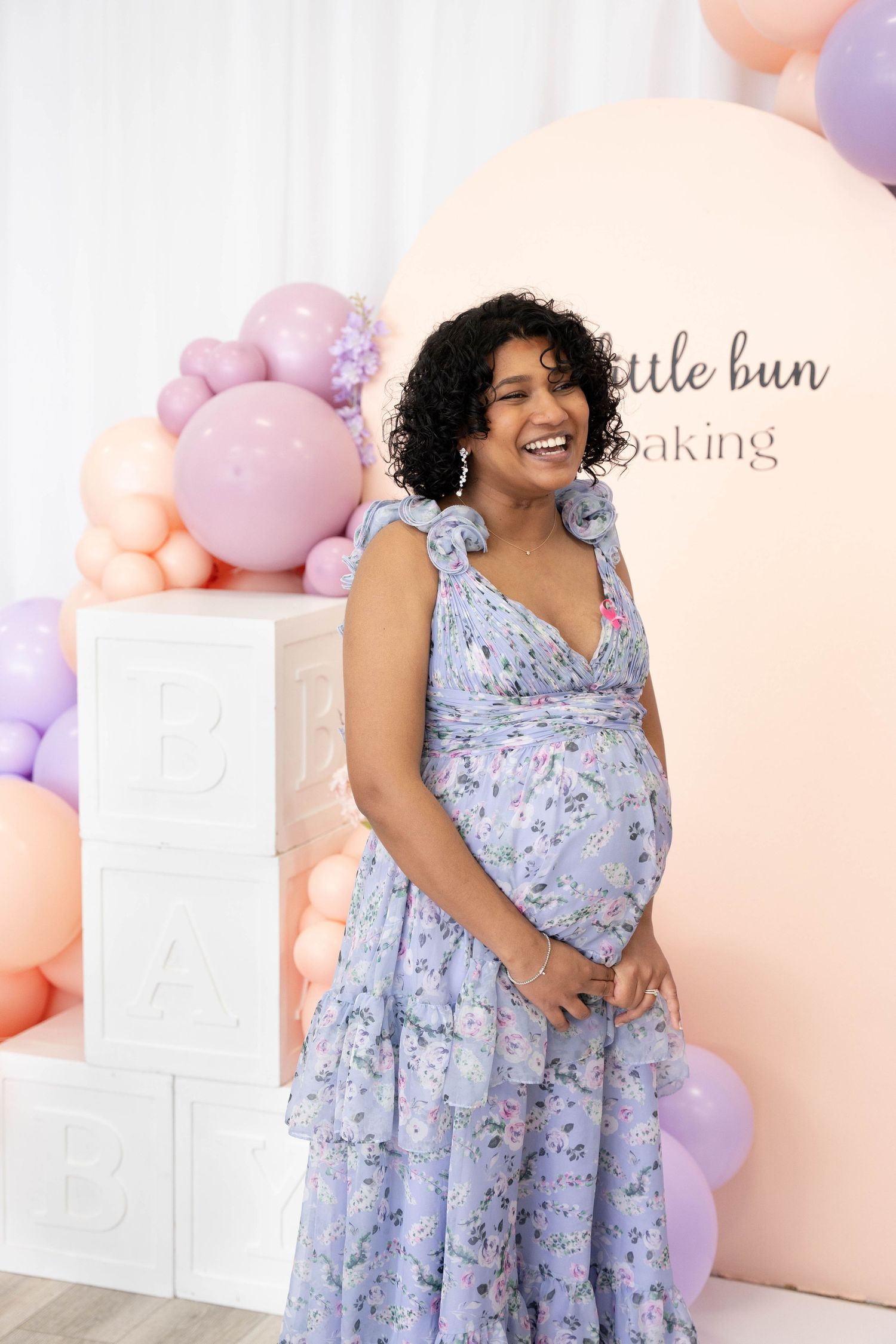 Laughing pregnant woman in a lavender floral dress stands by BABY letter blocks and a pastel balloon garland at her baby shower.