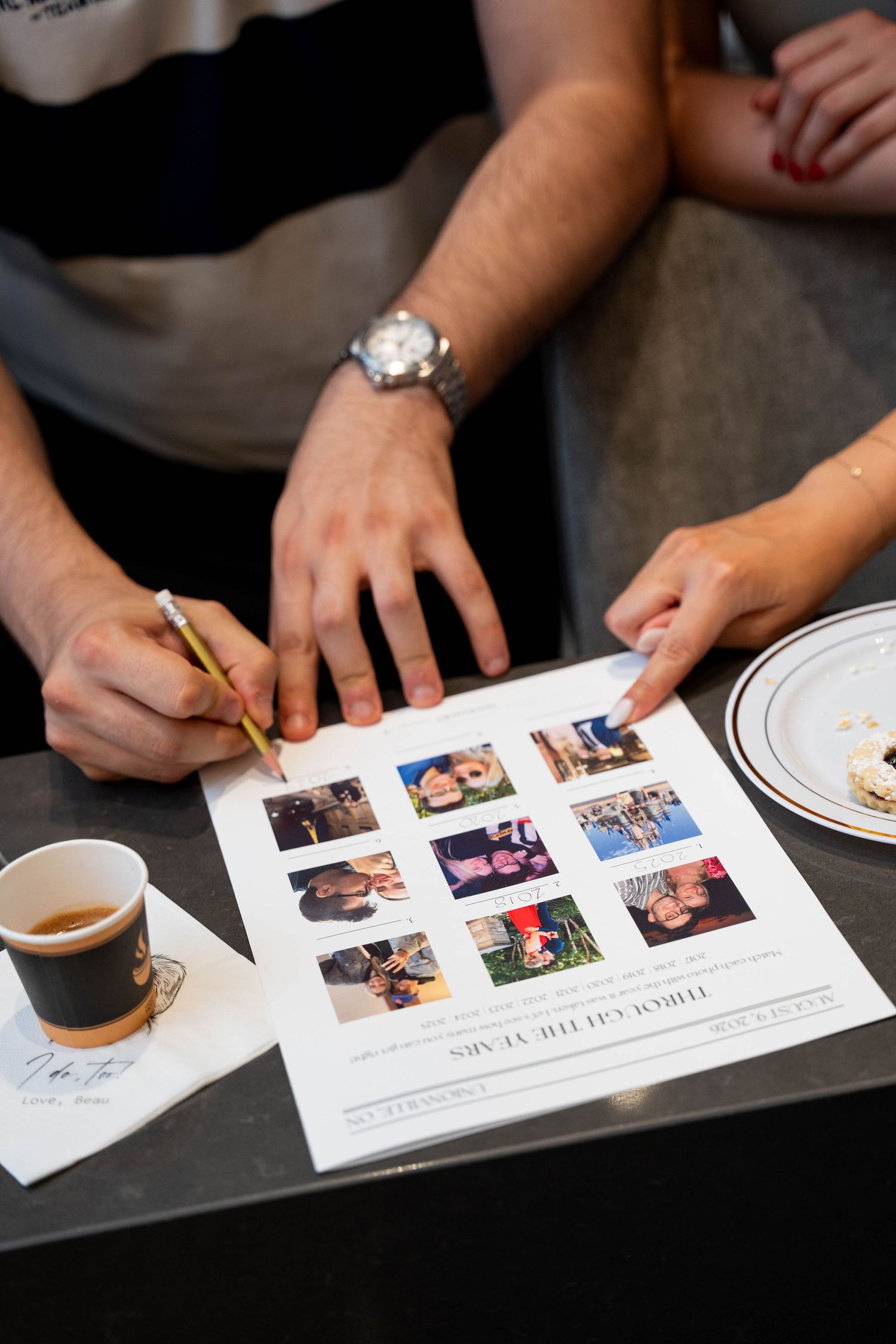 Close-up of a guest holding a pencil over a Through the Years photo game sheet beside a coffee cup at an engagement party.