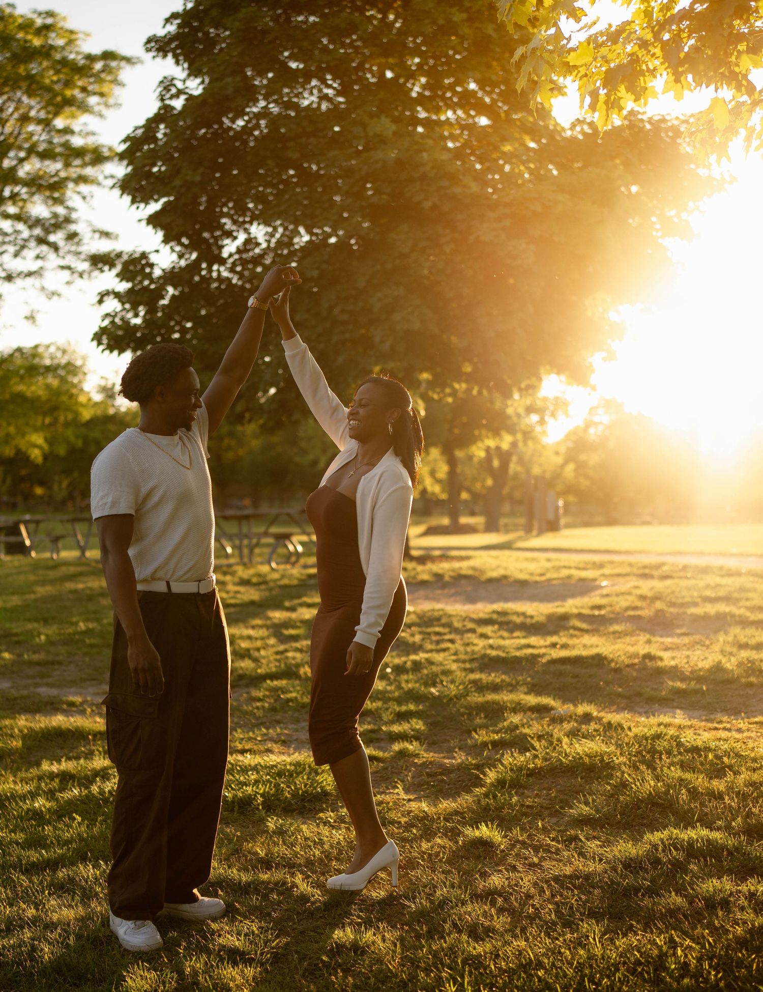 Couple dancing in a sunlit park at golden hour as the man twirls the woman beneath large shady trees.