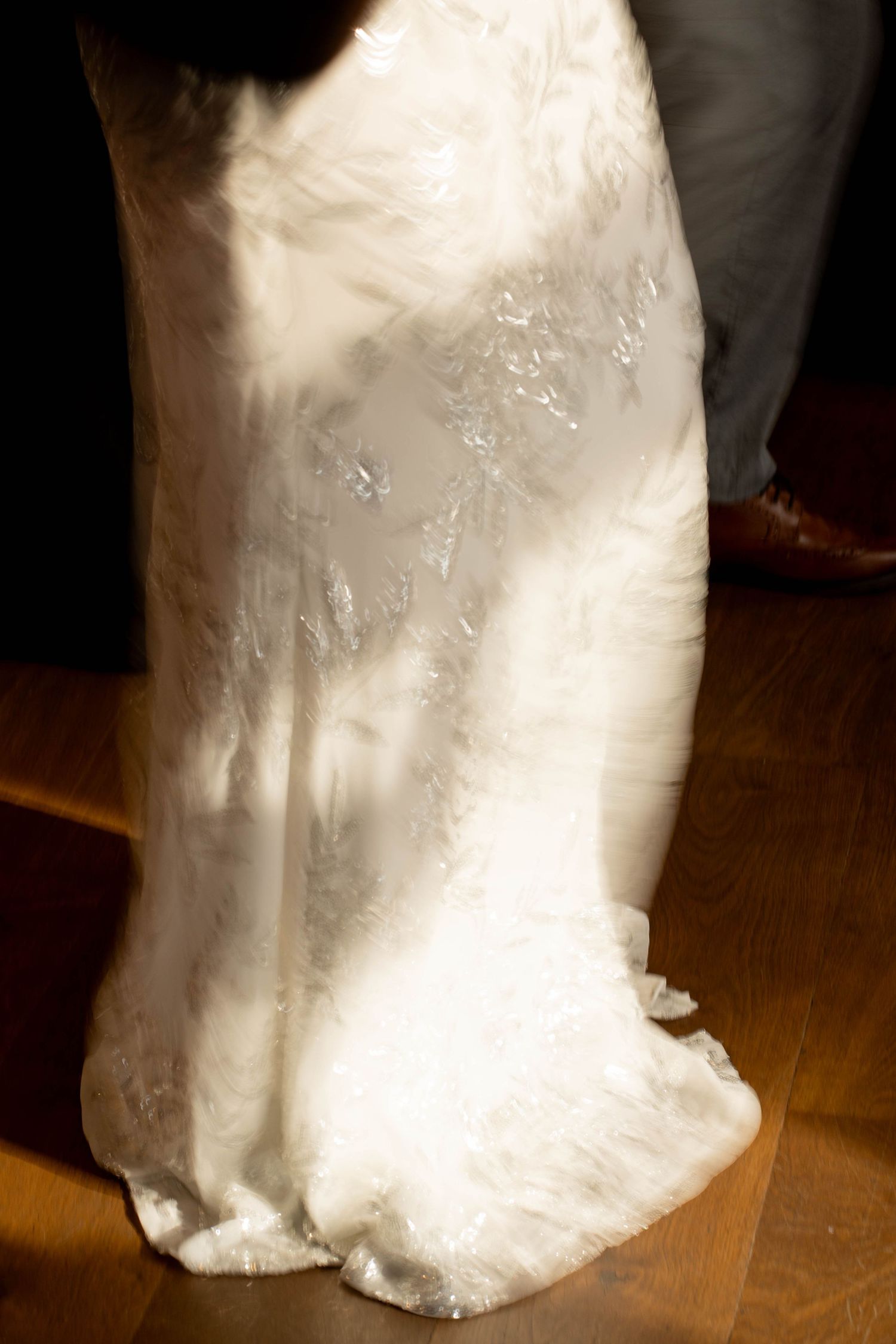 Close-up of a white beaded lace wedding gown train catching warm sunlight on a hardwood floor beside a groom's brown dress shoe.