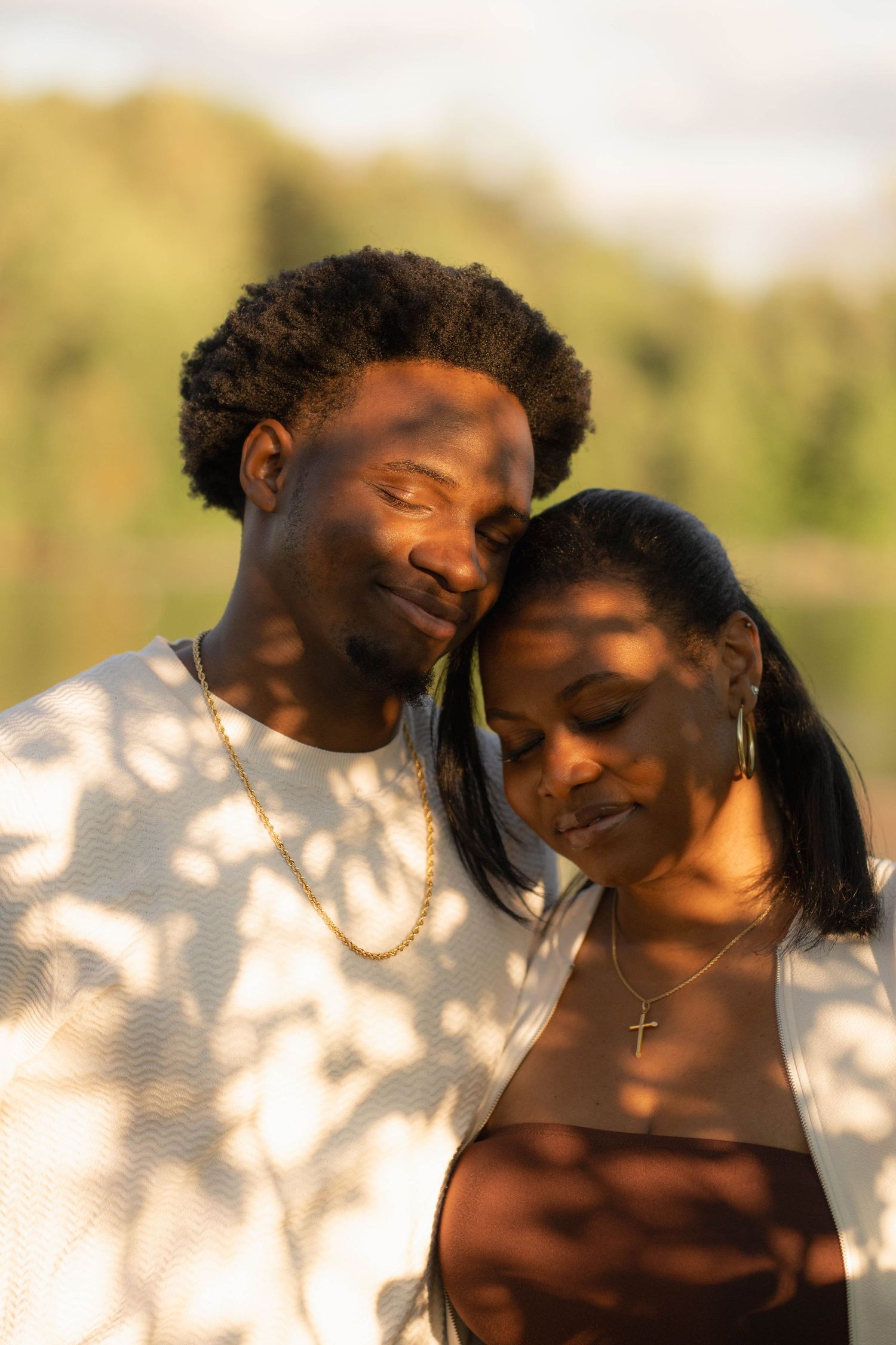 Couple embracing tenderly with eyes closed as the man rests his head on the woman's, dappled sunlight on their clothes.