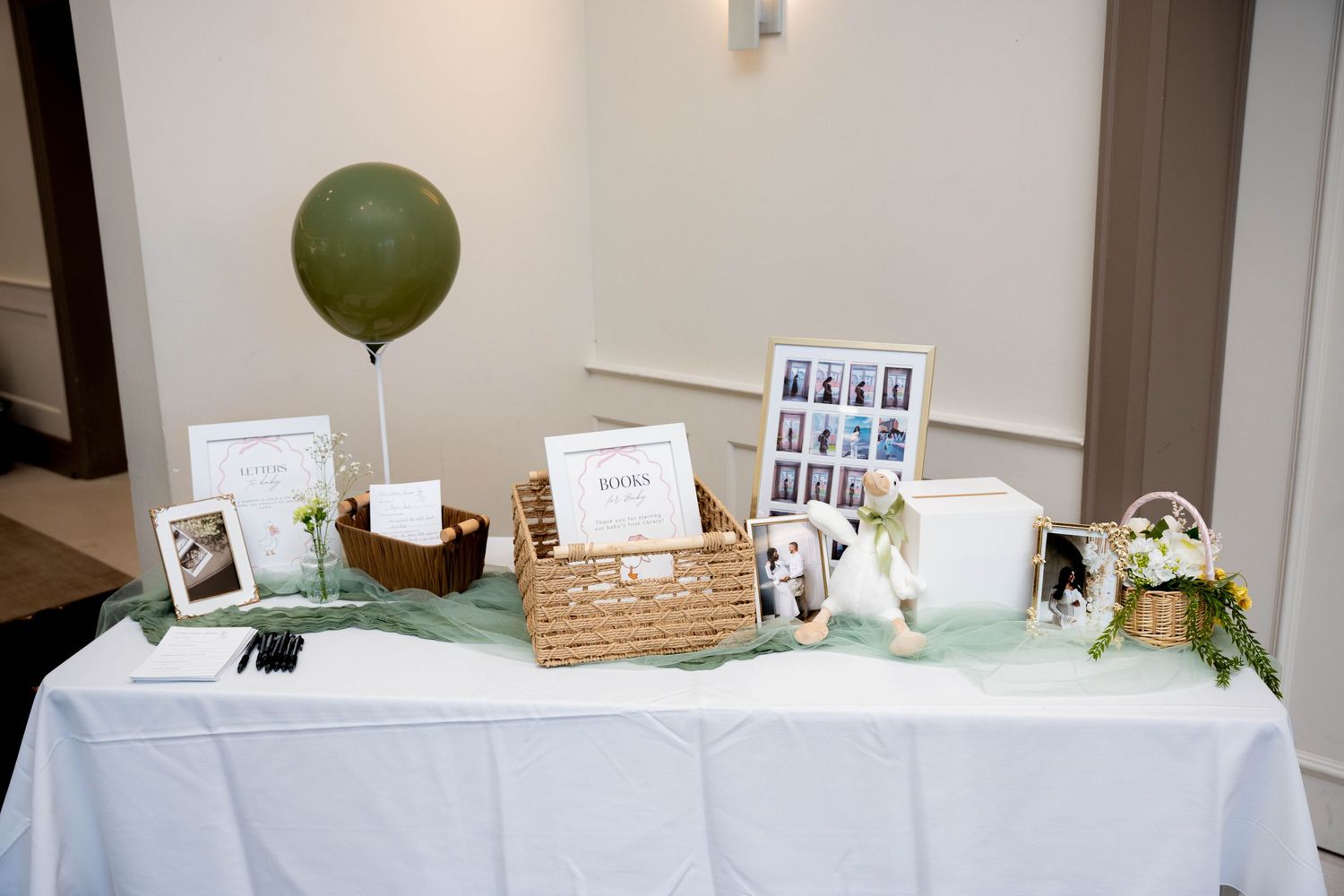 A baby shower gift table with white linens holds a basket, framed photos, a green balloon, and floral decor against a cream wall.