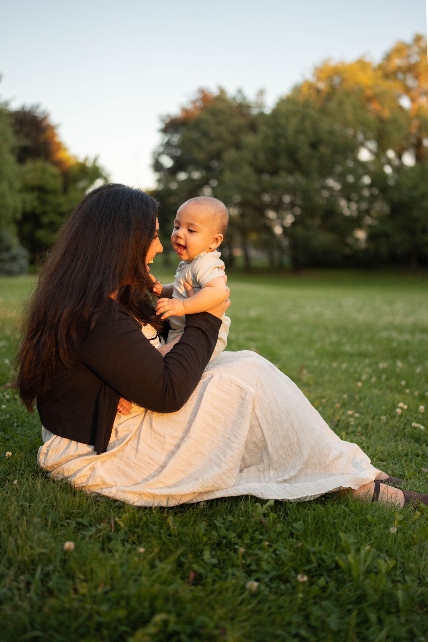 A mother in a white skirt and black cardigan sits on the grass and smiles at her laughing baby in a park at sunset.