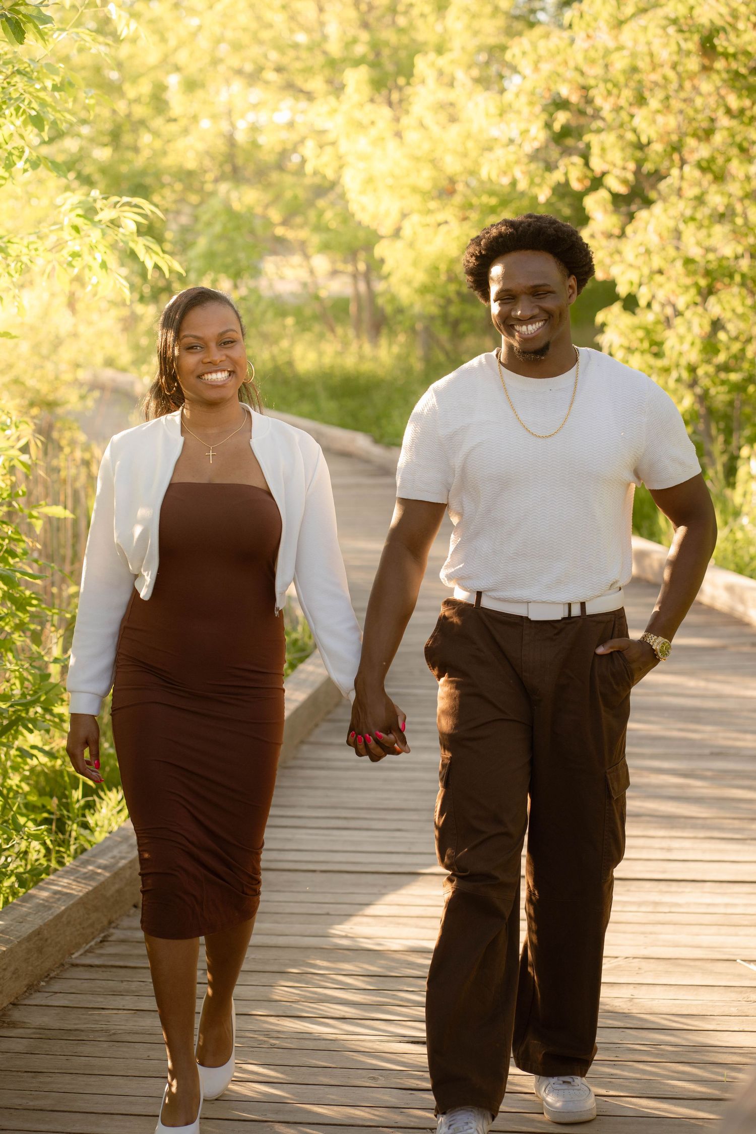 Joyful couple in white and brown outfits walking hand in hand down a sunlit wooden boardwalk through trees.