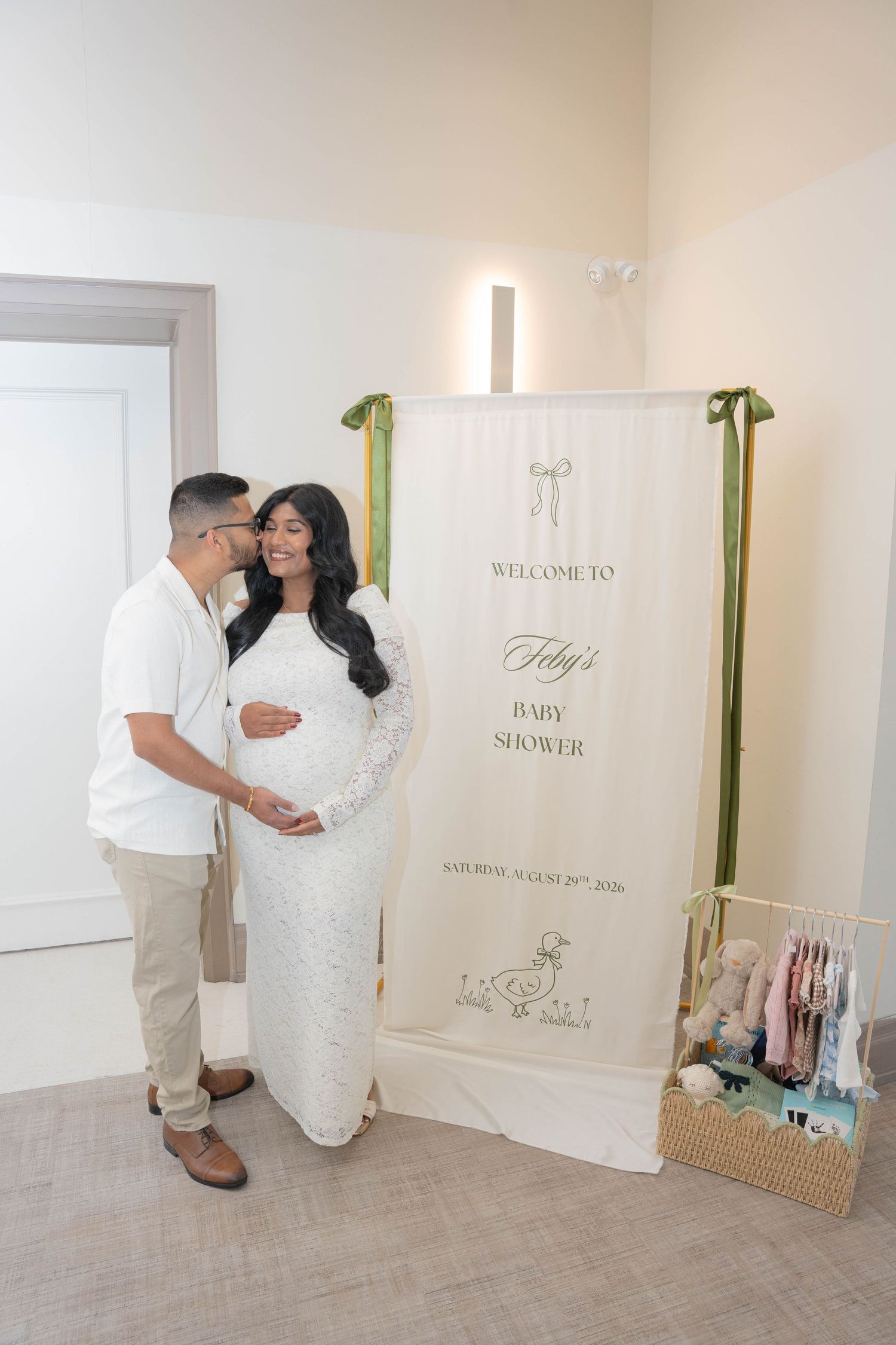 An expecting couple embraces beside a baby shower welcome sign, the mother-to-be in a white lace gown and a basket of plush toys nearby.