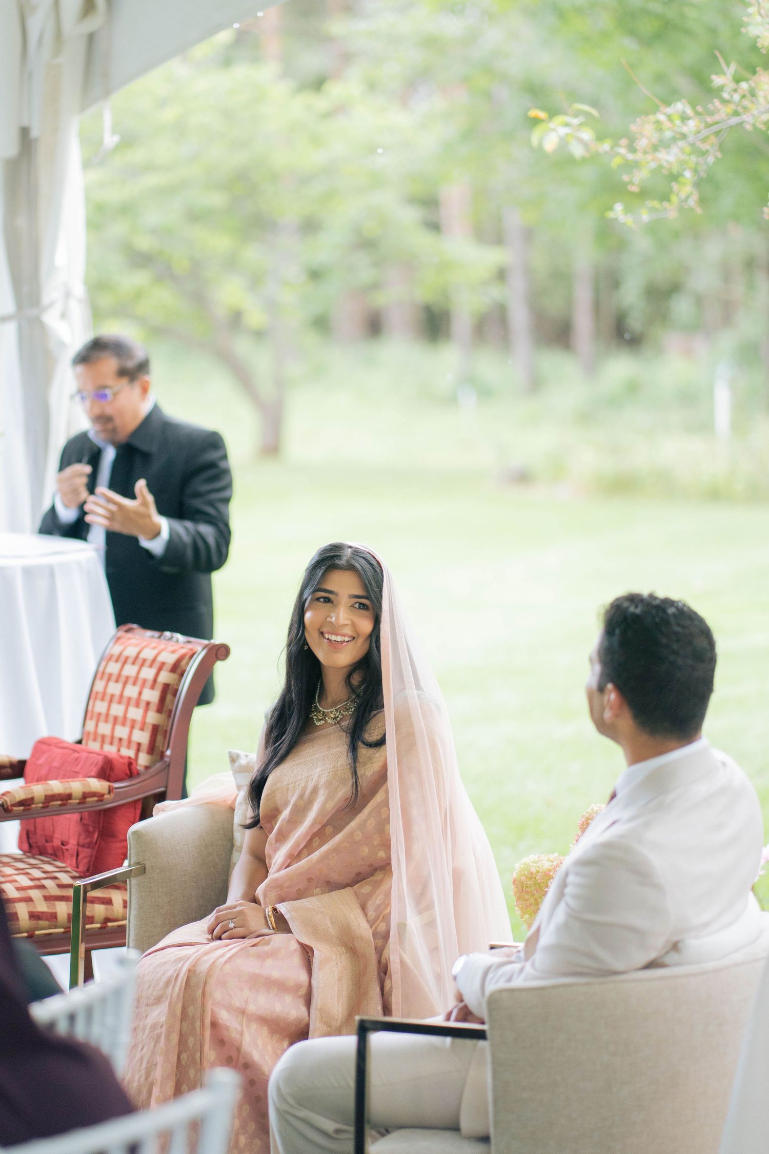 A smiling bride in a peach sari and veil sits beside her groom while a man in a suit speaks into a microphone at an outdoor tent ceremony.