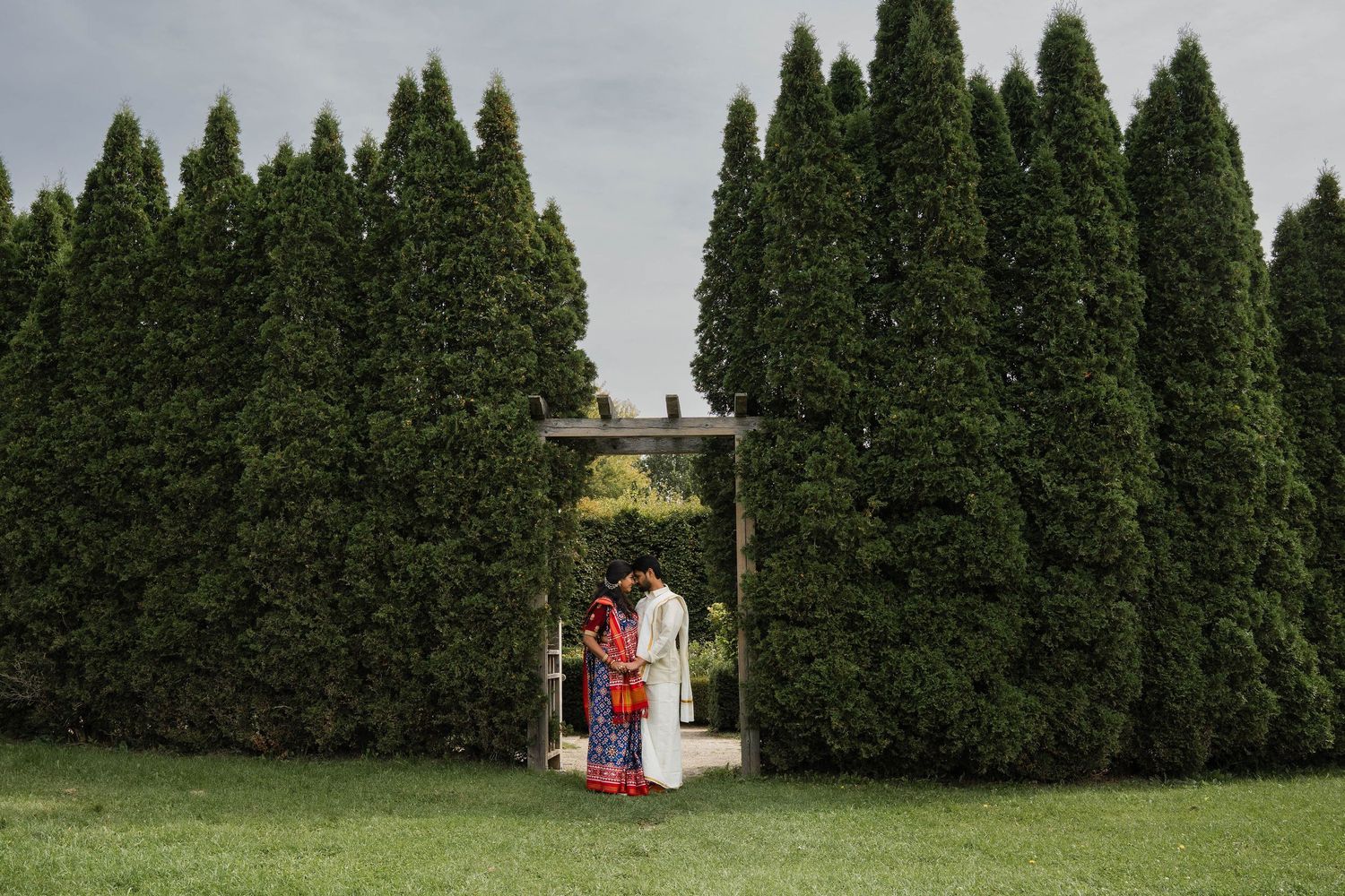A couple in traditional Indian attire stands beneath a small wooden arbor in front of tall evergreen trees on a green lawn.