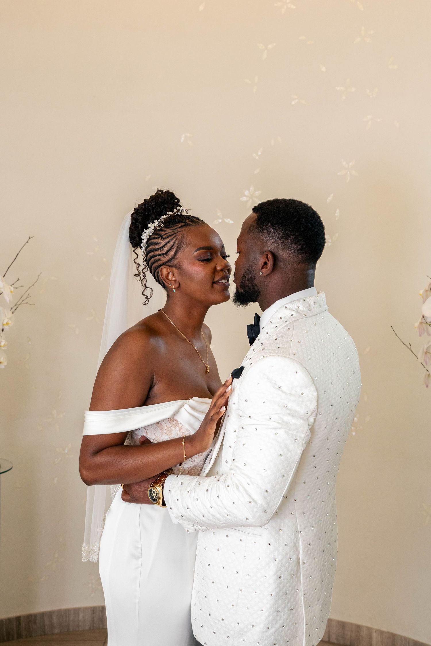 Bride in an off-shoulder white gown and veil embraces her groom in a white beaded tuxedo jacket and black bow tie.
