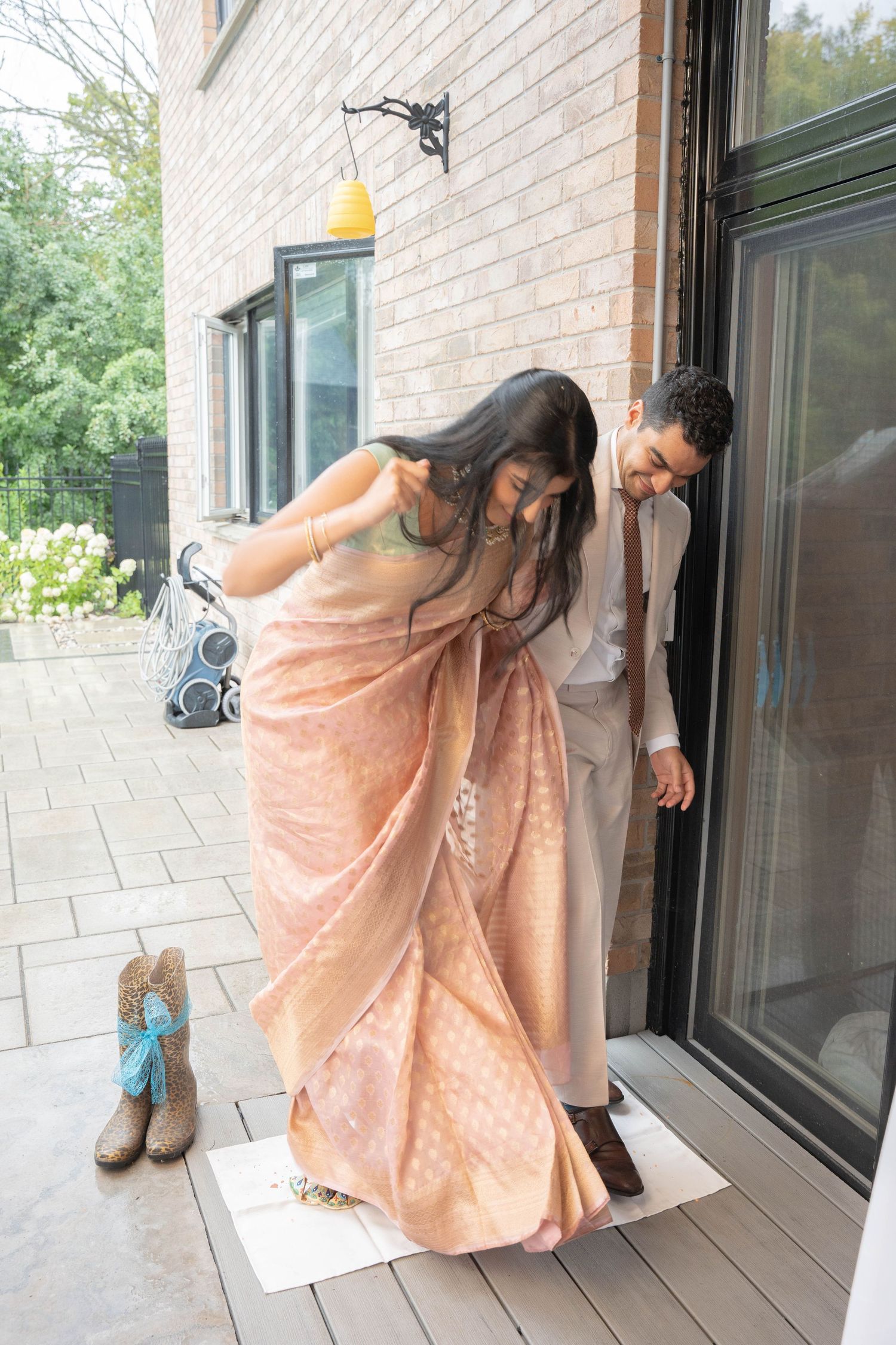 A woman in a peach silk saree and a man in a beige suit bend toward a white mat in a playful wedding ritual.