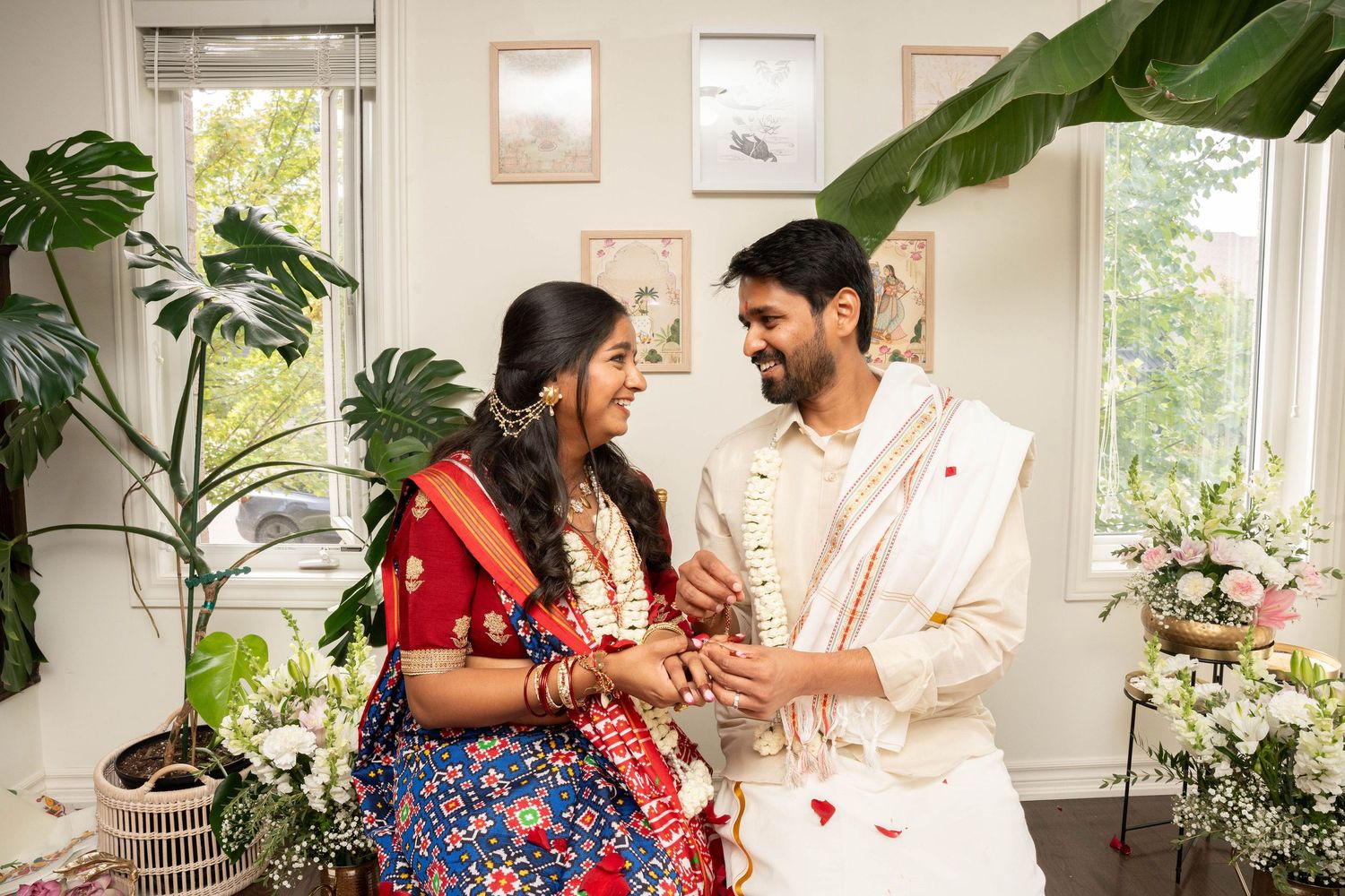 A smiling couple in traditional Indian wedding clothes exchanges rings in a bright room decorated with fresh flowers and green plants.