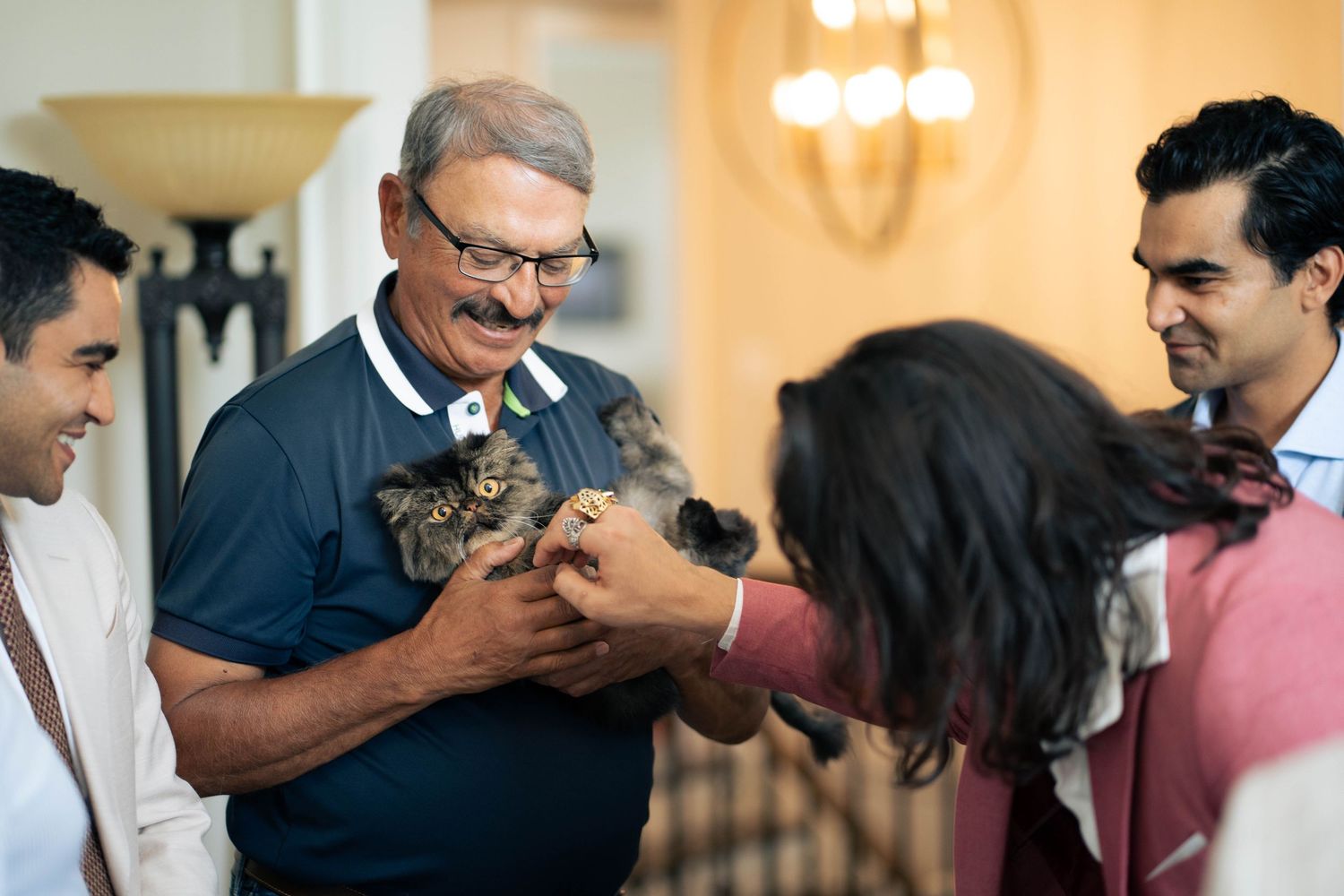 Smiling older man in glasses holds a small kitten as a woman and two men look on with delight indoors.