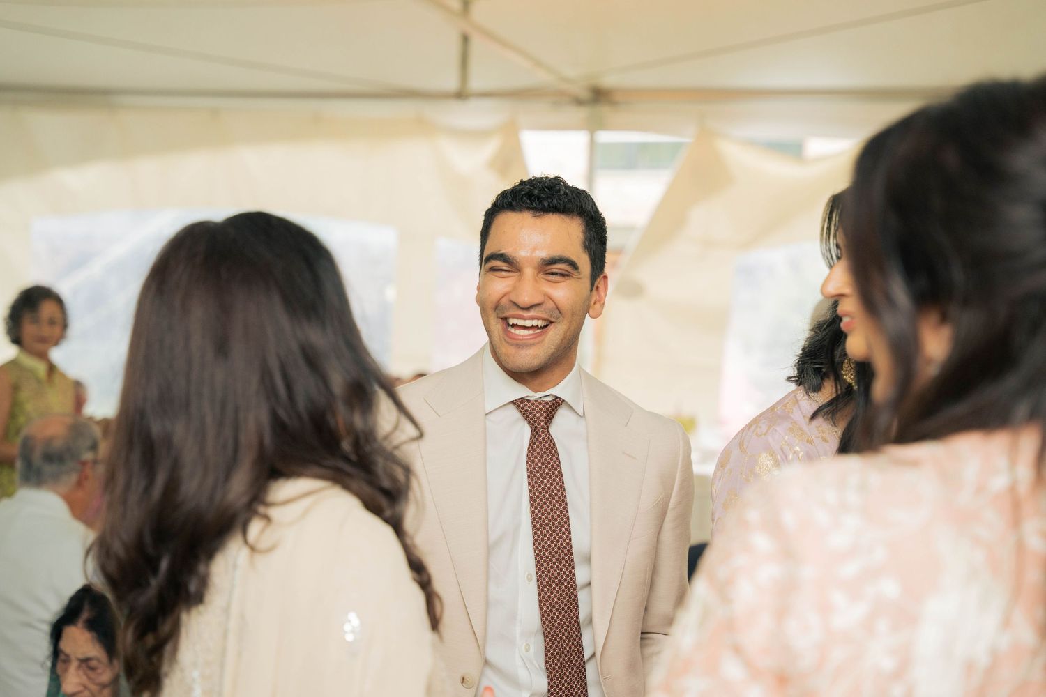 A smiling groom in a cream suit and brown tie laughs while mingling with wedding guests under a white tent.