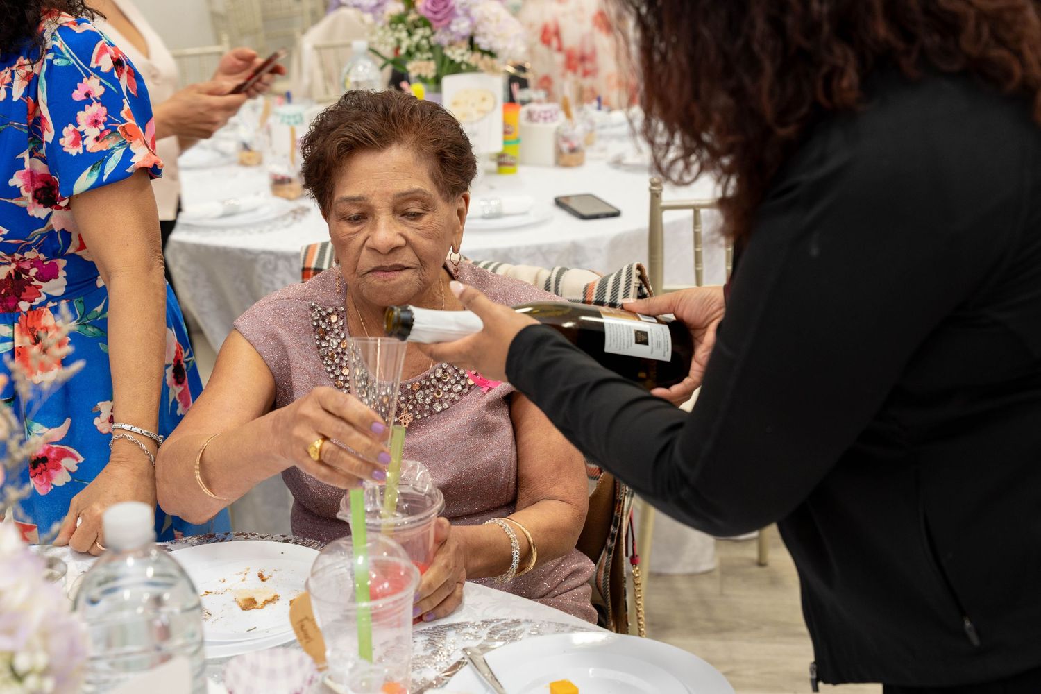 A woman in a pink top smiles as a guest pours sparkling wine into her glass at a decorated baby shower table.