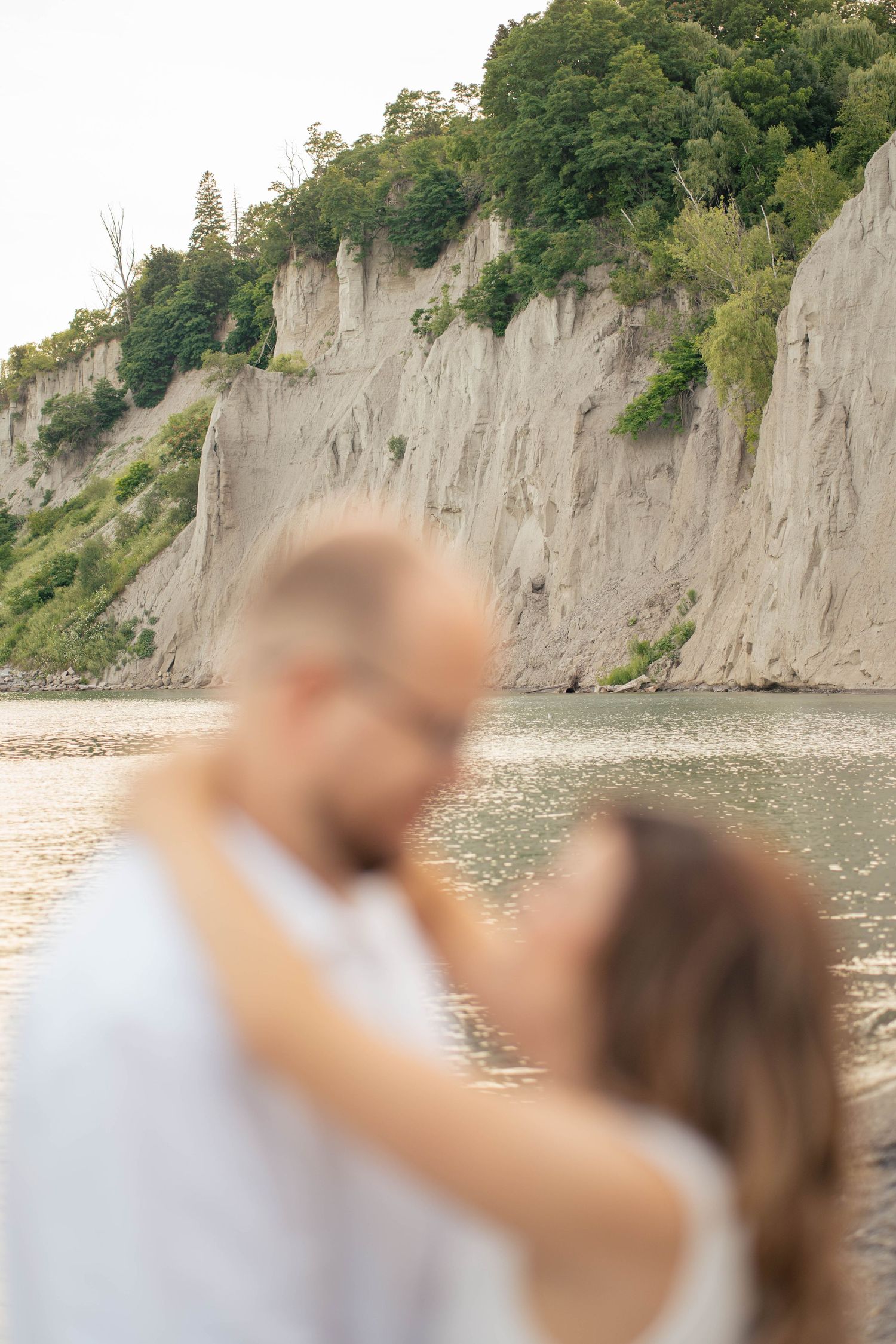 Blurred couple embracing on a lakeshore beach with tall eroded cliffs and green trees rising in the background.
