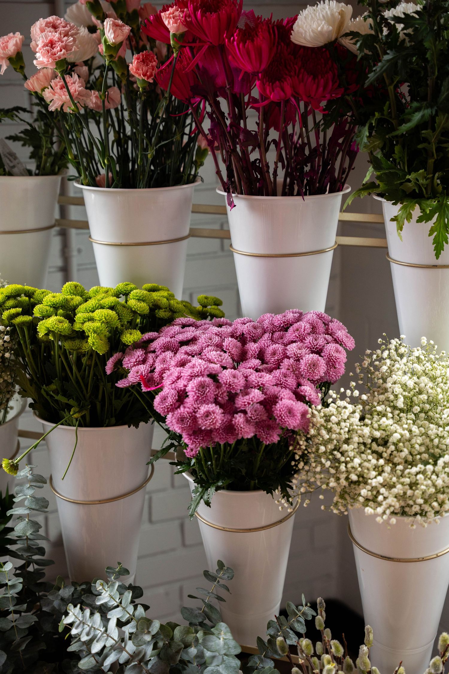 White buckets display fresh pink and red carnations, green chrysanthemums, pink mums and baby's breath alongside eucalyptus greenery.