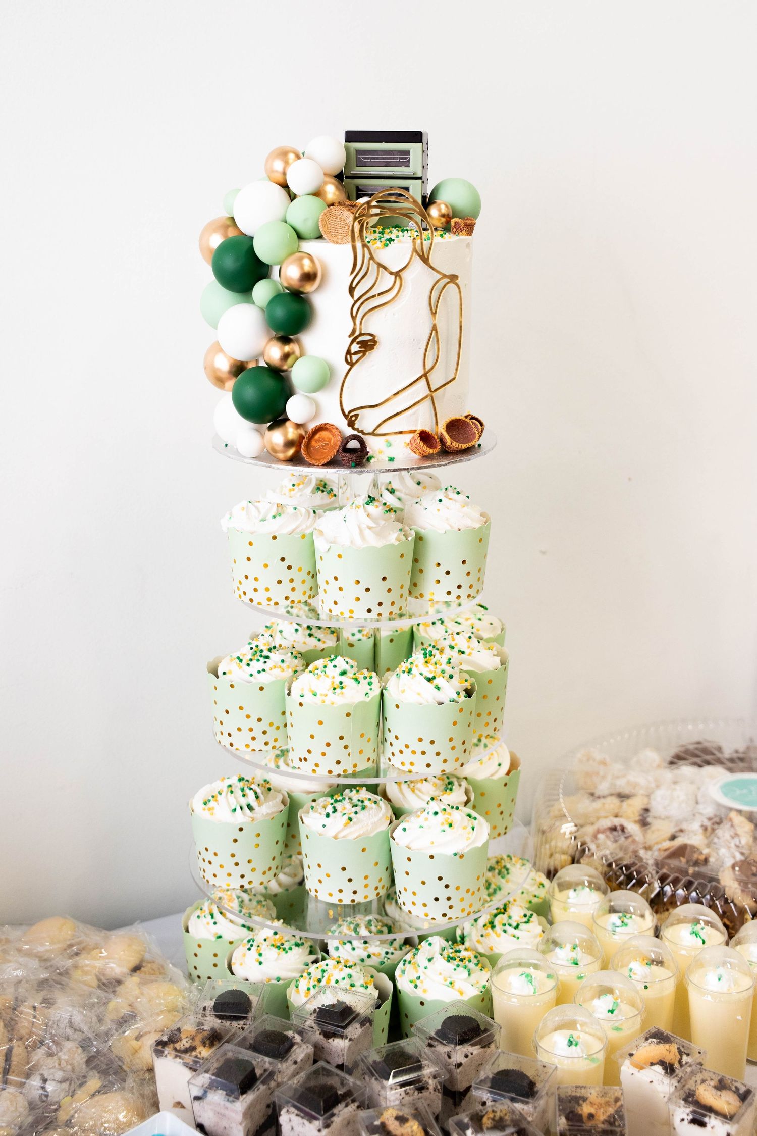 Green and white dessert tower with sprinkle-topped cupcakes, mini desserts and a cake decorated with gold, green and white spheres.