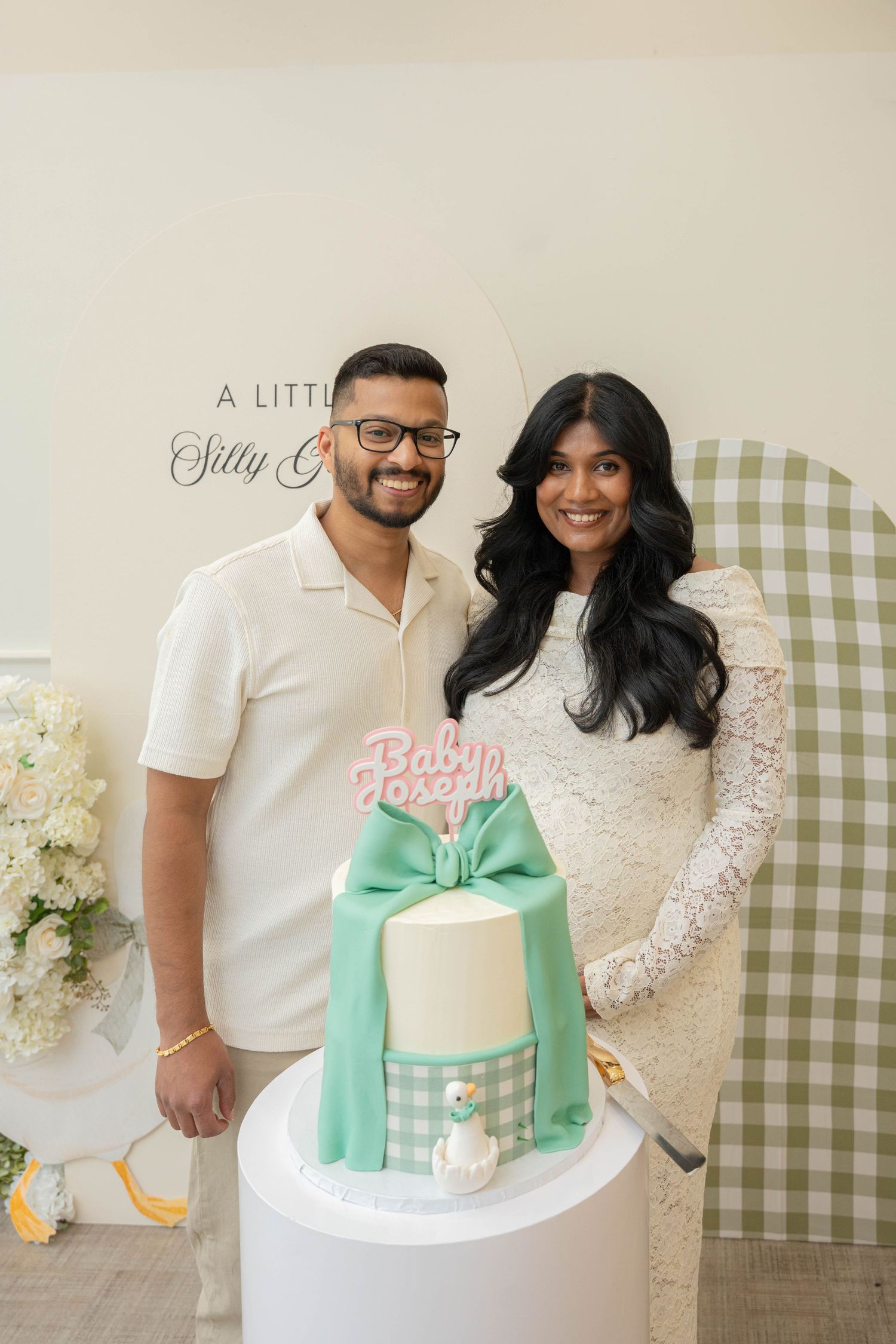 Smiling couple poses beside a mint green bow cake topped with a Baby Joseph sign at a gingham-themed baby shower.