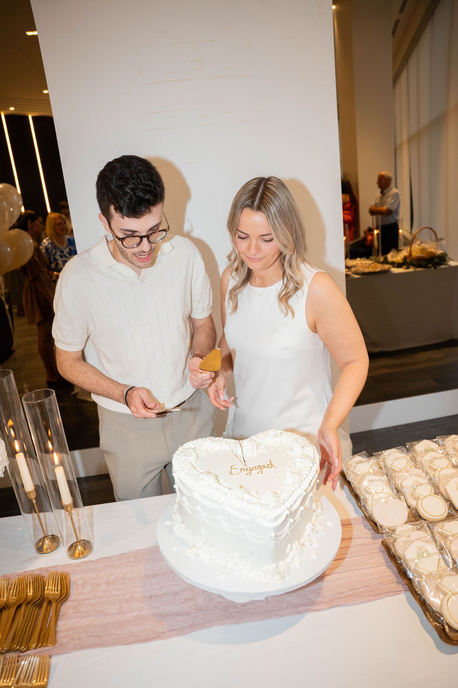A smiling couple prepares to cut their heart-shaped engagement cake at a dessert table during their engagement party celebration.