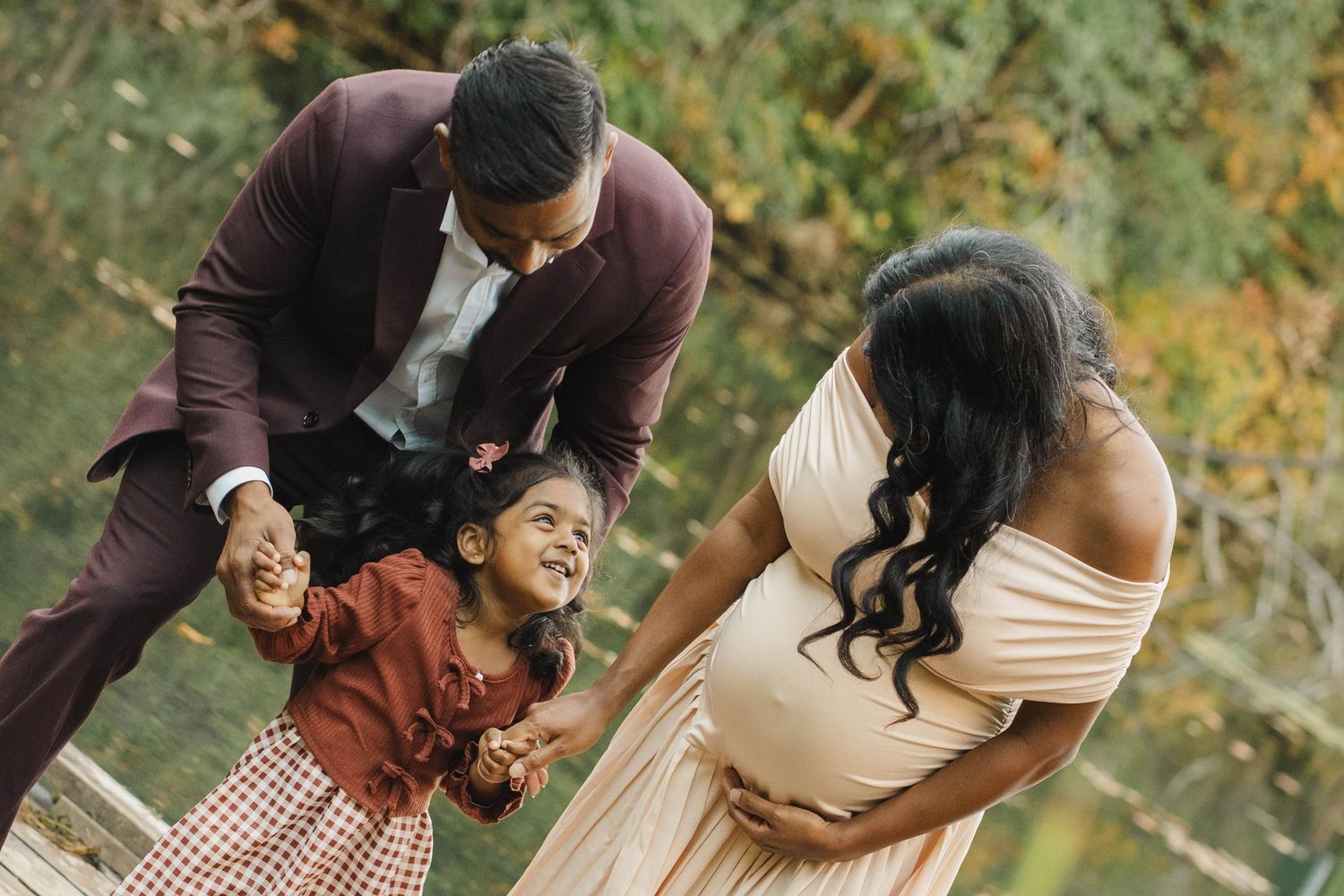 Smiling little girl holds her father's hands while her pregnant mother in a blush gown looks on during an outdoor maternity photo session.