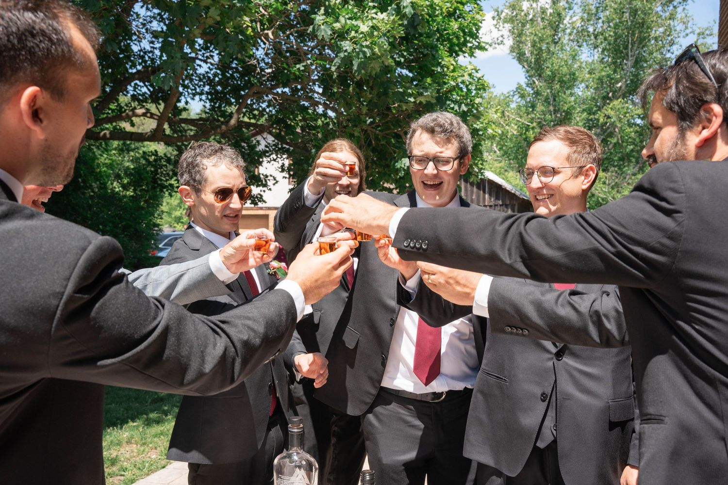 A group of groomsmen in suits raise small glasses in a cheerful toast outdoors on a sunny day.