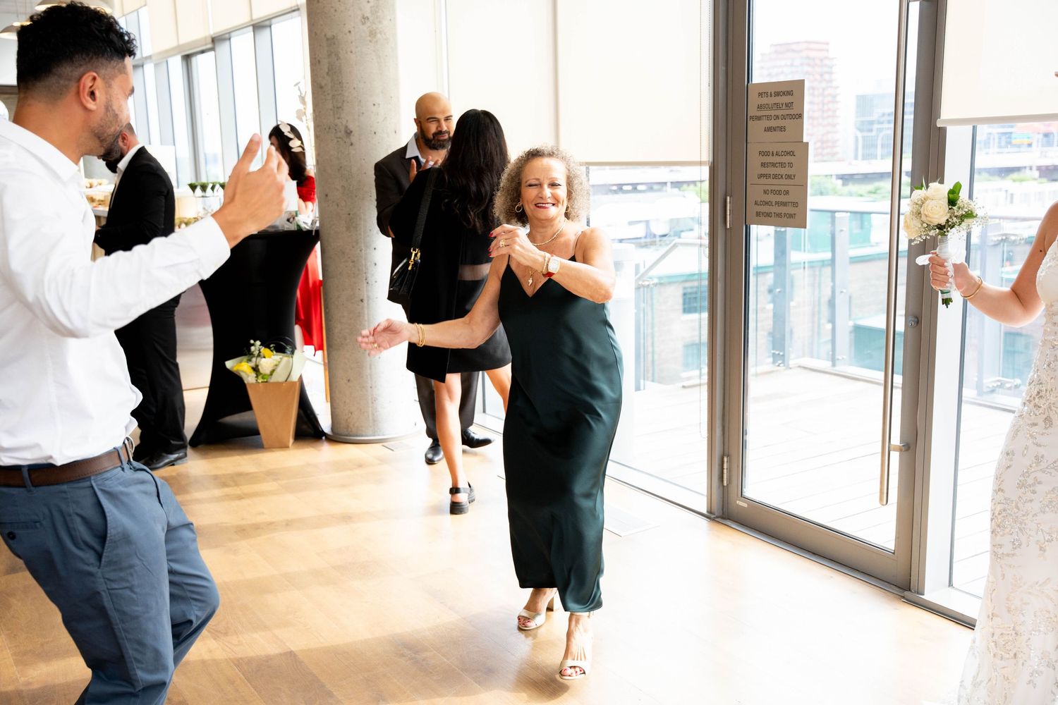 Wedding guests dance and celebrate on a wooden floor in a bright room with large windows as the bride holds her bouquet.