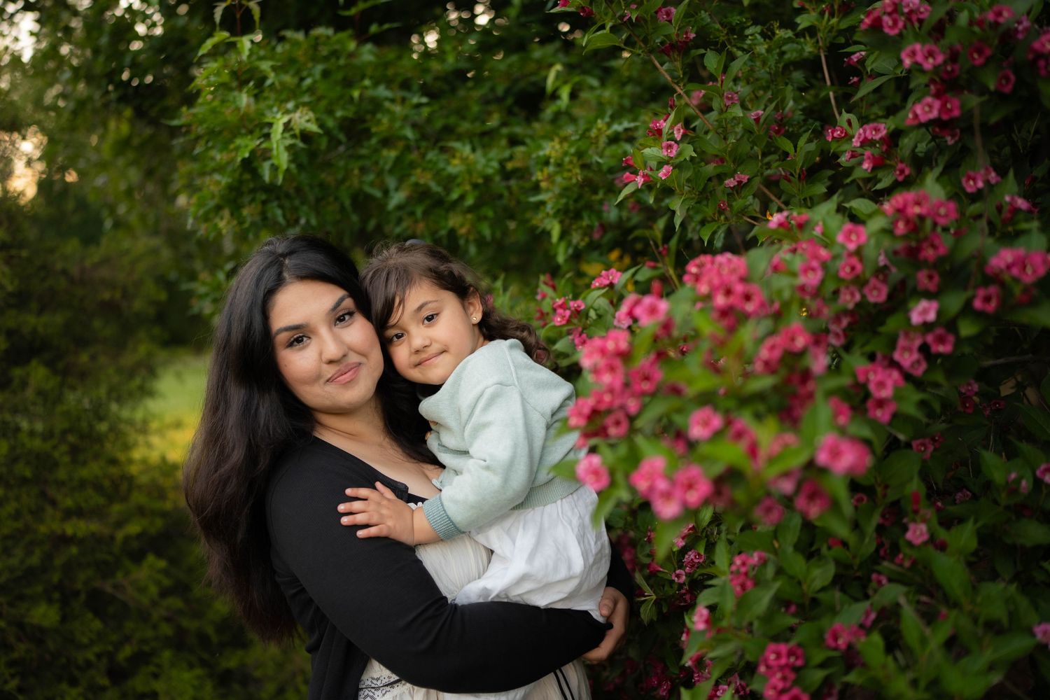 A smiling mother in black holds her young daughter in a mint cardigan beside a blooming pink weigela bush in a garden.