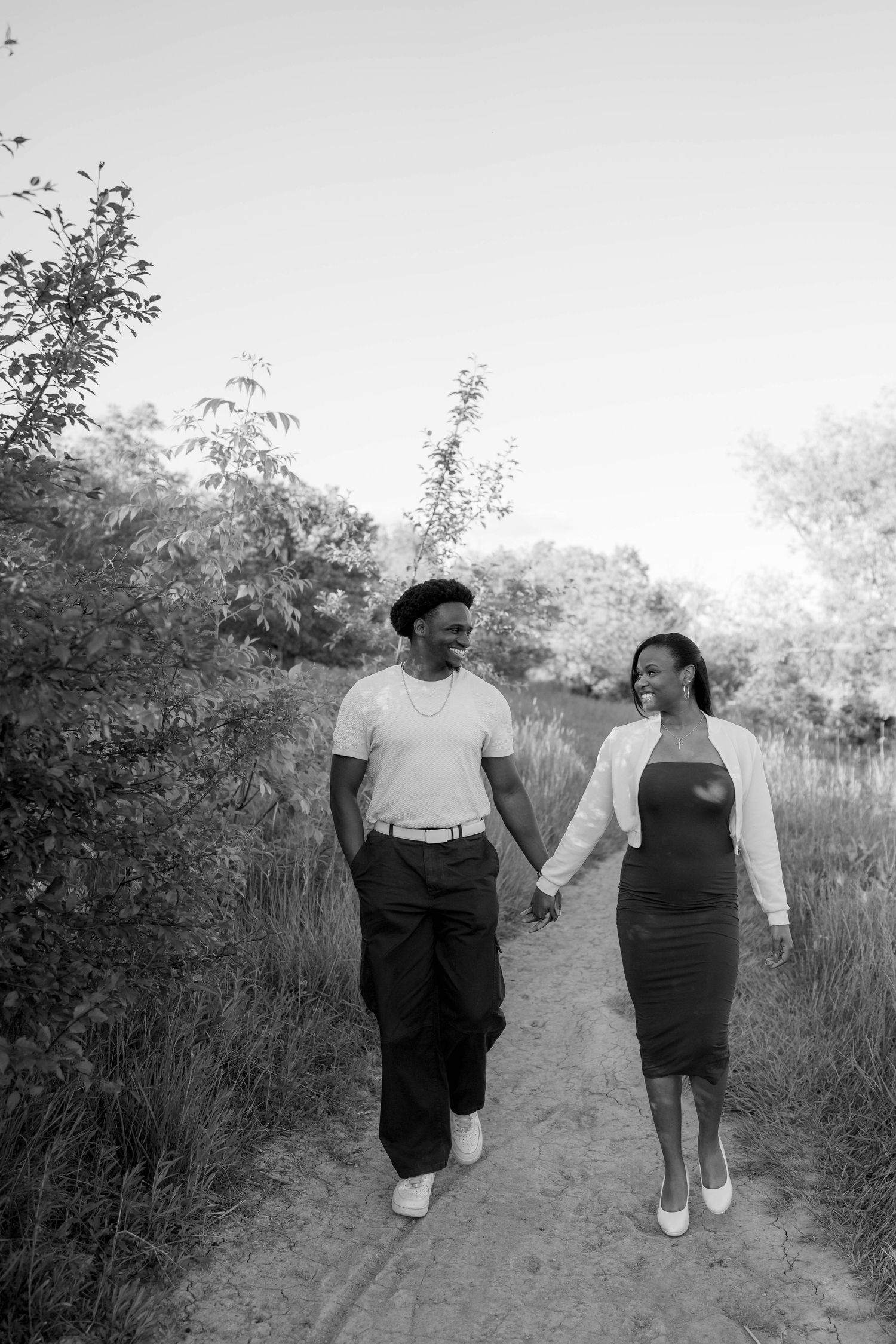 Black and white photo of a smiling engaged couple holding hands and walking along a grassy outdoor trail.
