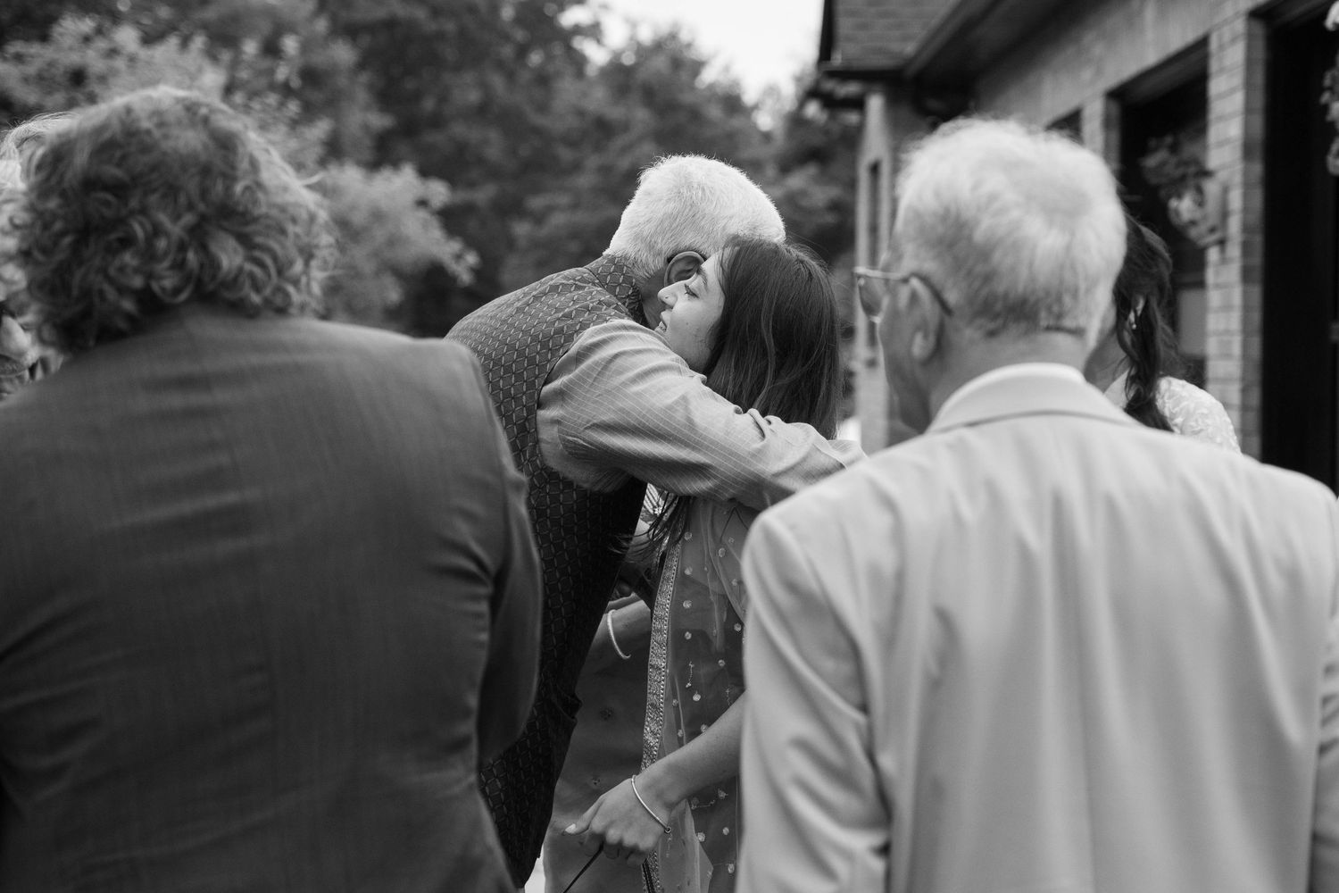 Black and white photo of an elderly man hugging a woman warmly as guests stand nearby outside a stone building.