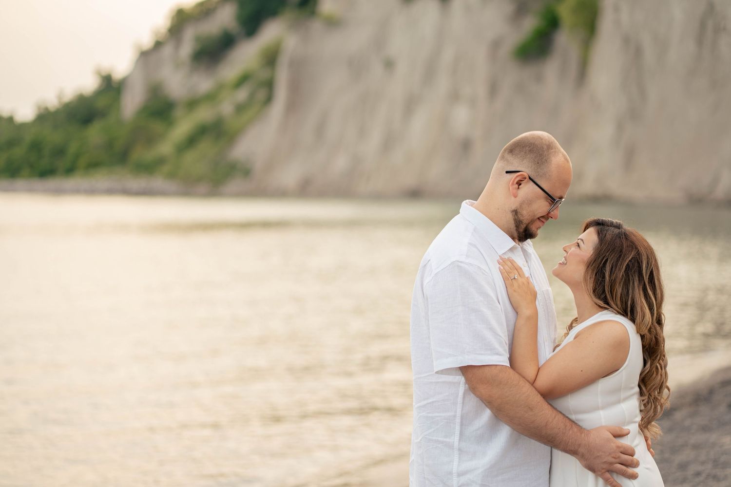 Couple embracing and smiling on a beach at sunset with tall cliffs and calm water behind them.