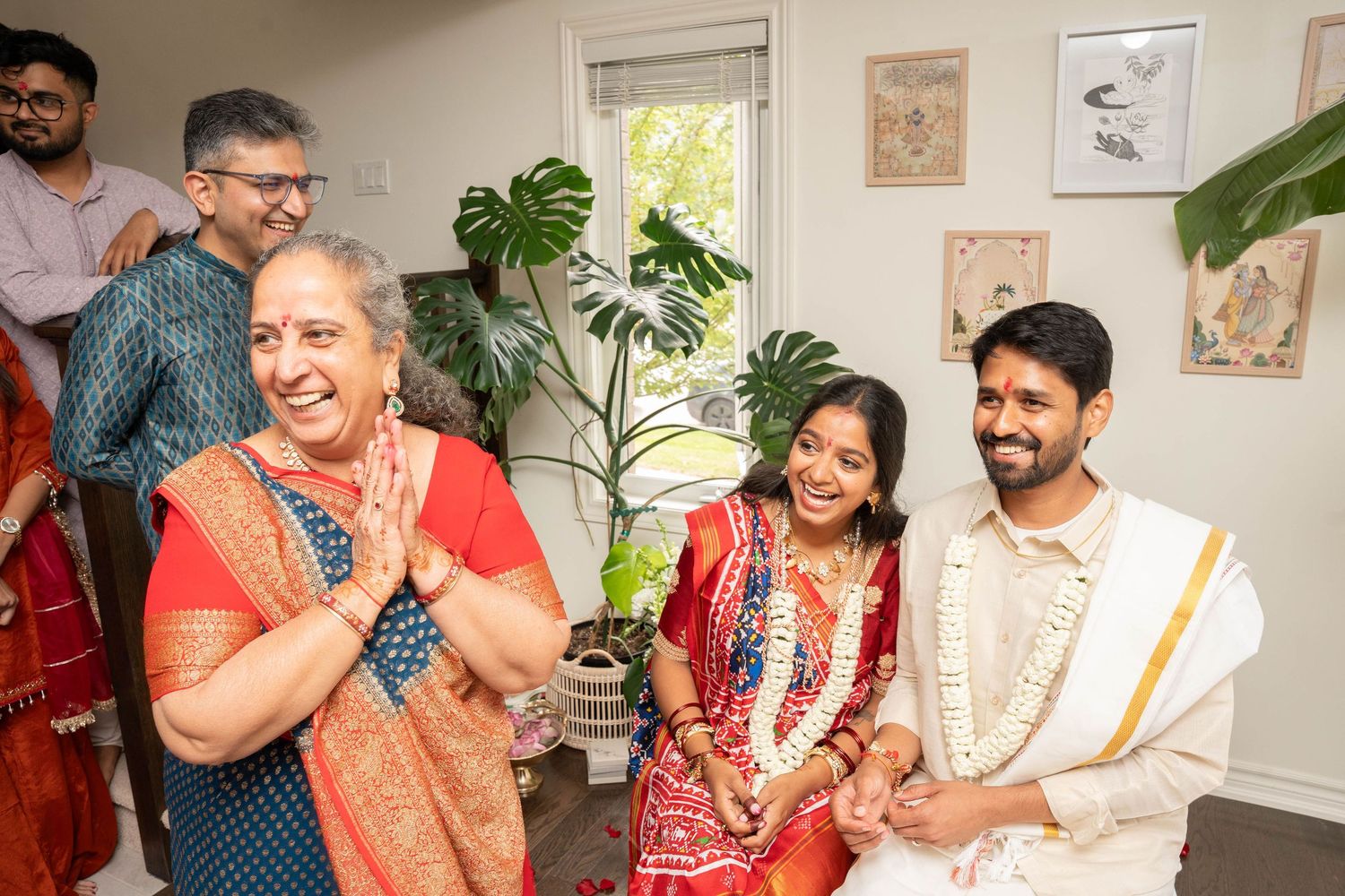 Smiling woman in an orange and red sari with hands pressed together beside a seated couple during an indoor Indian wedding ceremony.
